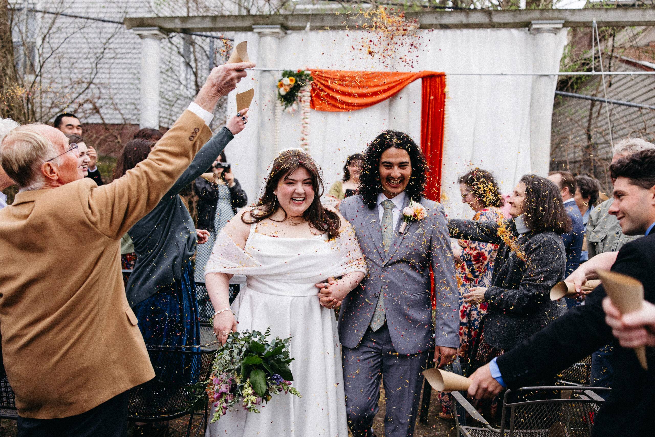 A newlywed couple walks hand in hand, smiling broadly as guests throw colorful confetti in a celebratory outdoor ceremony setting.