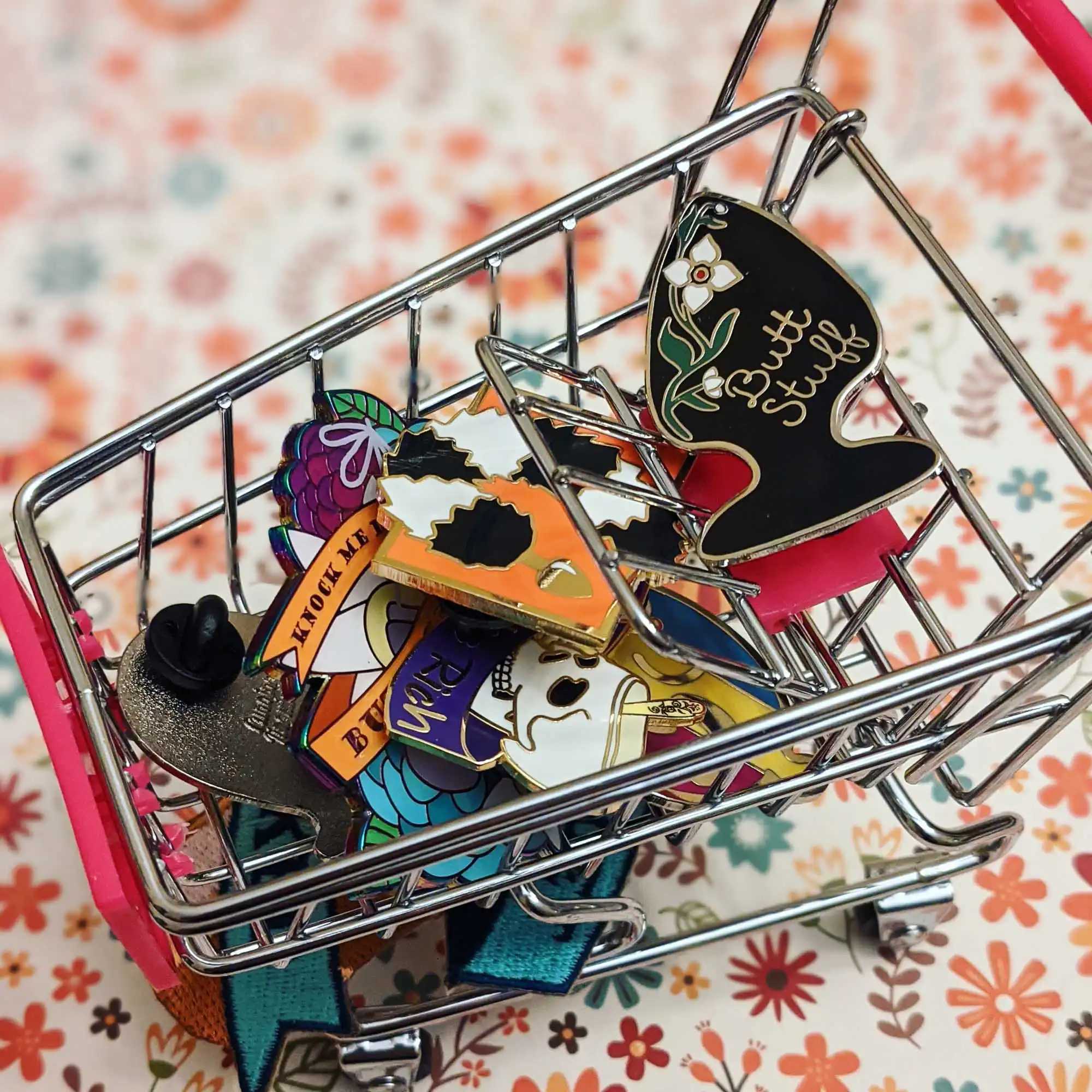 A mini shopping cart filled with enamel pins, including one shaped like a butt plug and one with a skull.