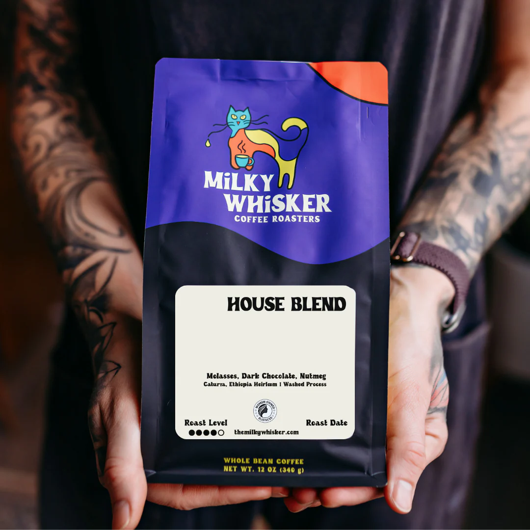 Hands holding a bag of The Milky Whisker House Blend coffee with a bold purple and yellow cat logo on the label.