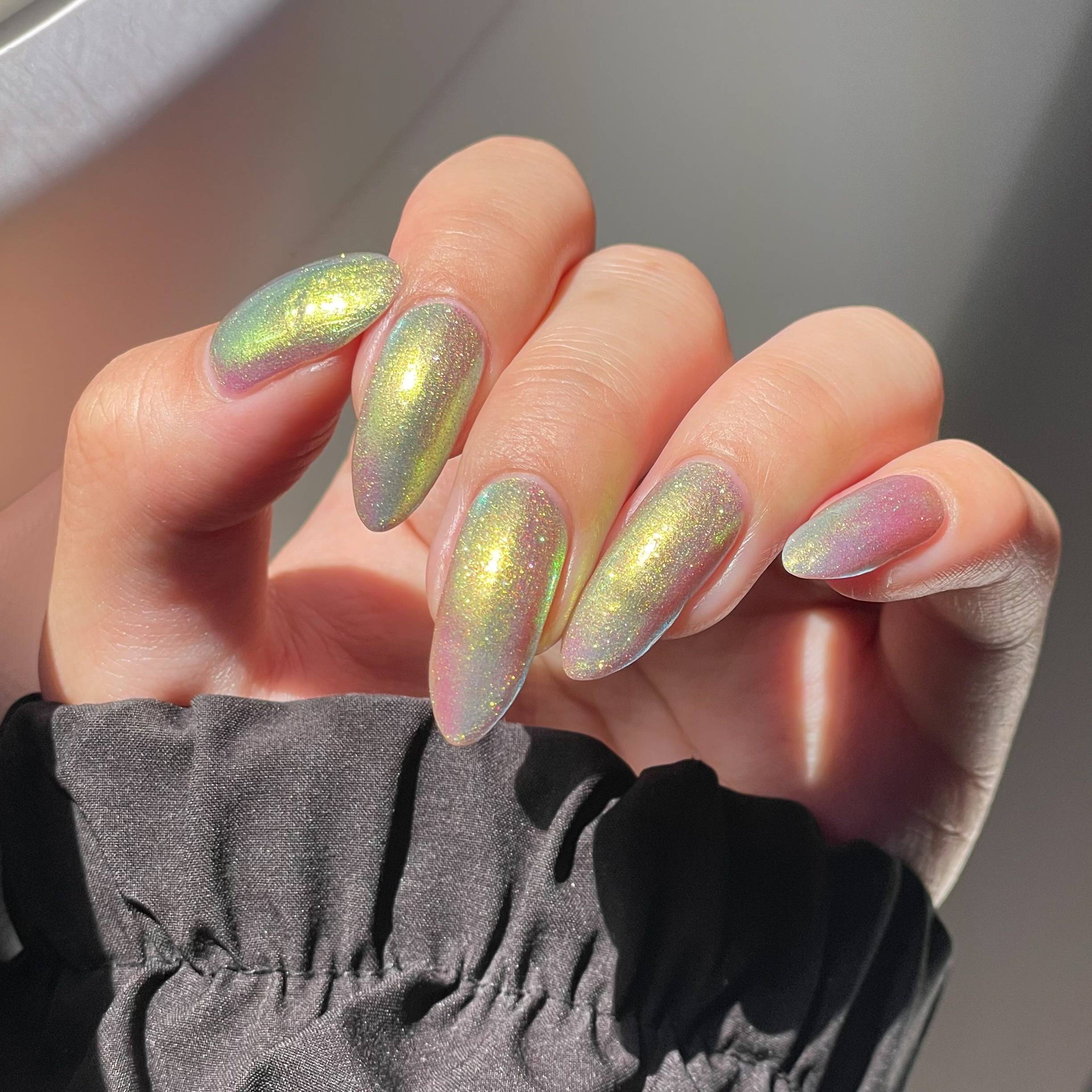 A pale skinned hand elegantly shows off a sparkly shifting green-gold-grey-pink nail polish on long almond nails.
