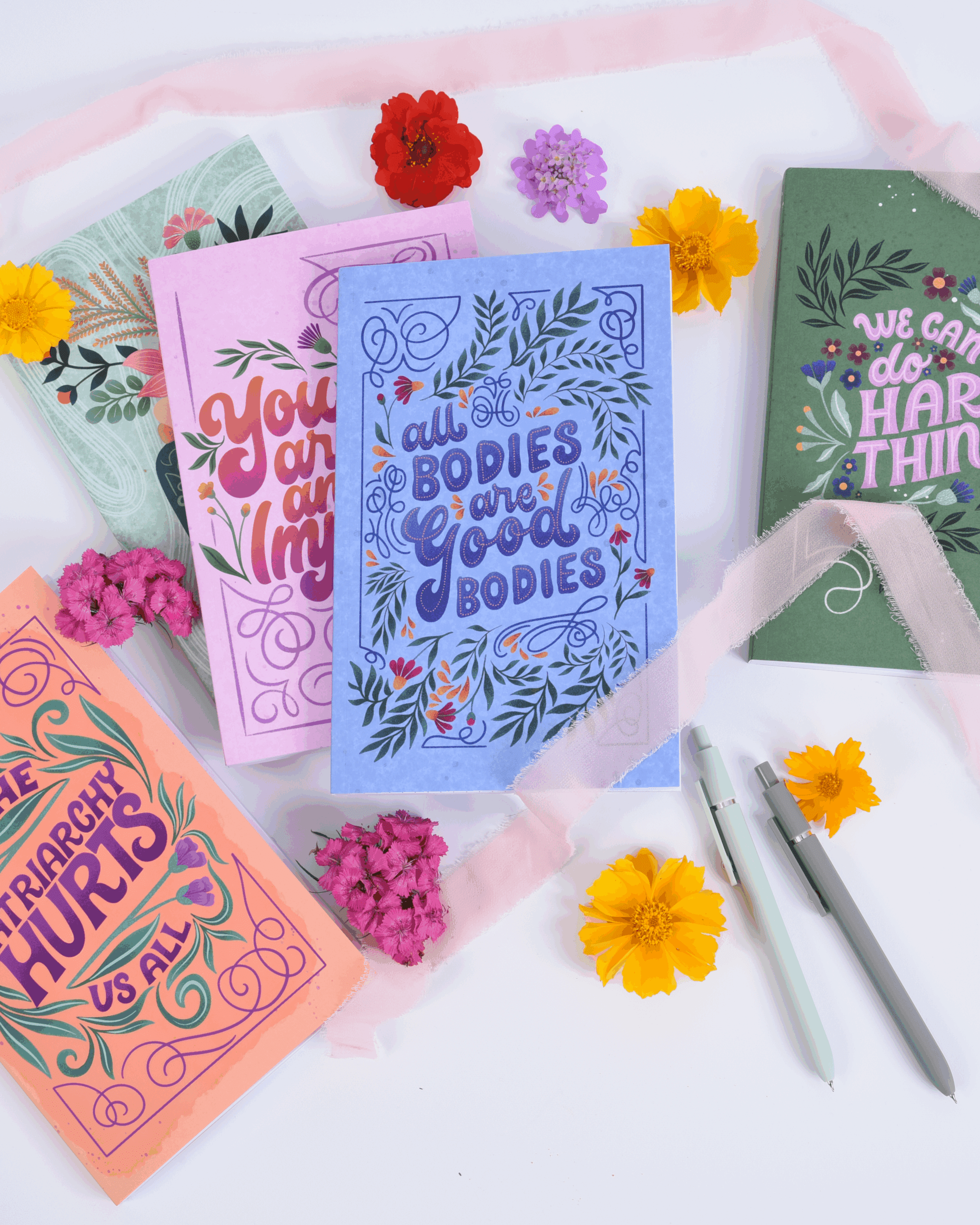 Several colorful notebooks on a white background ith flowers and ribbons. The notebooks all have positive messages
