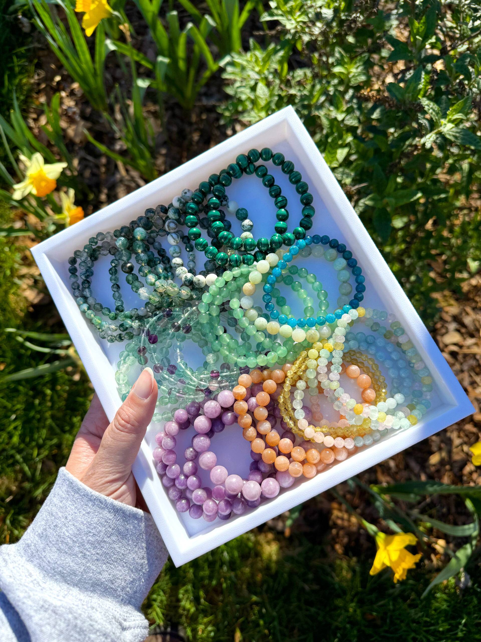 A hand holding a white tray filled with colorful crystal beaded bracelets with flowers in the backdrop.