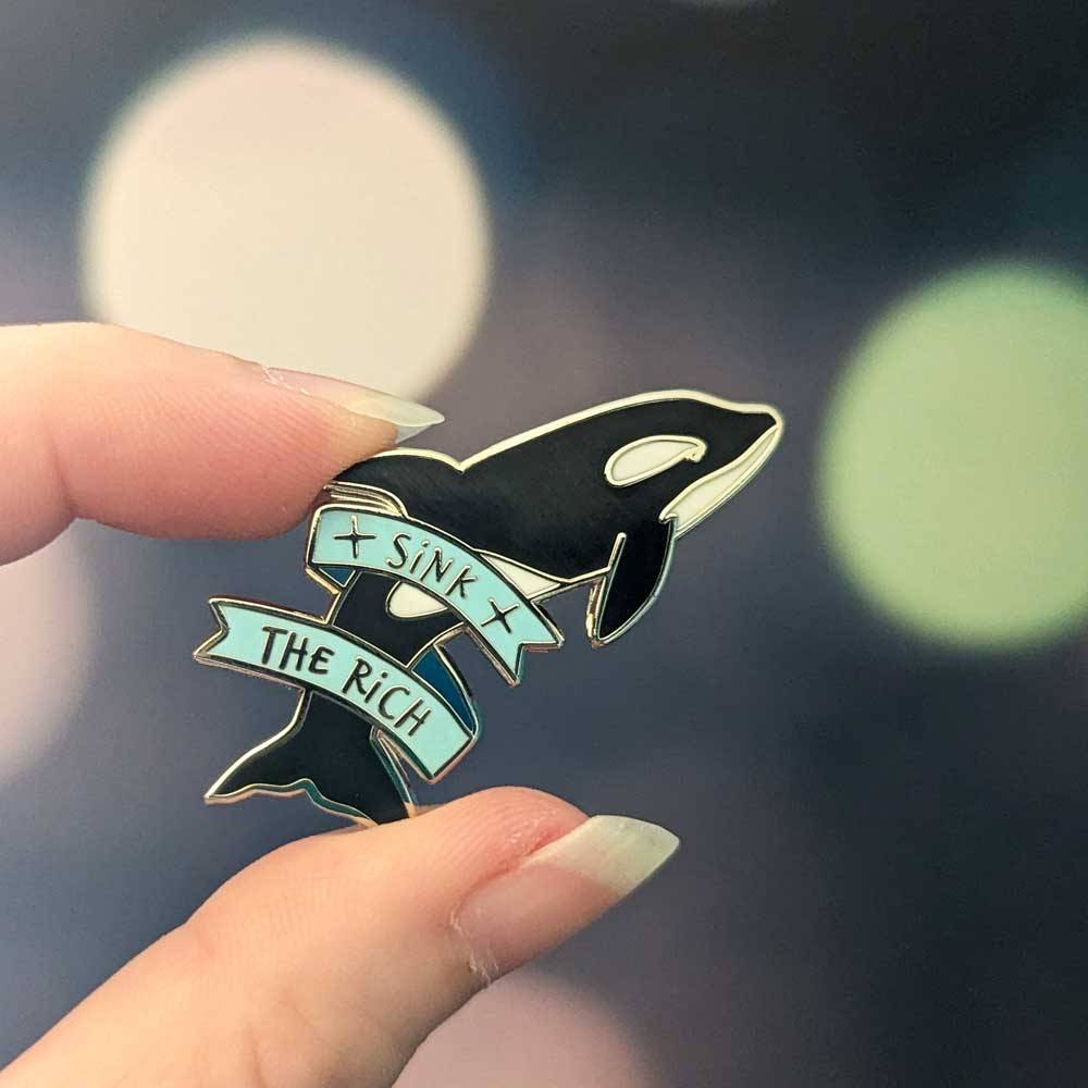 An enamel pin shaped like an orca; a ribbon has text 