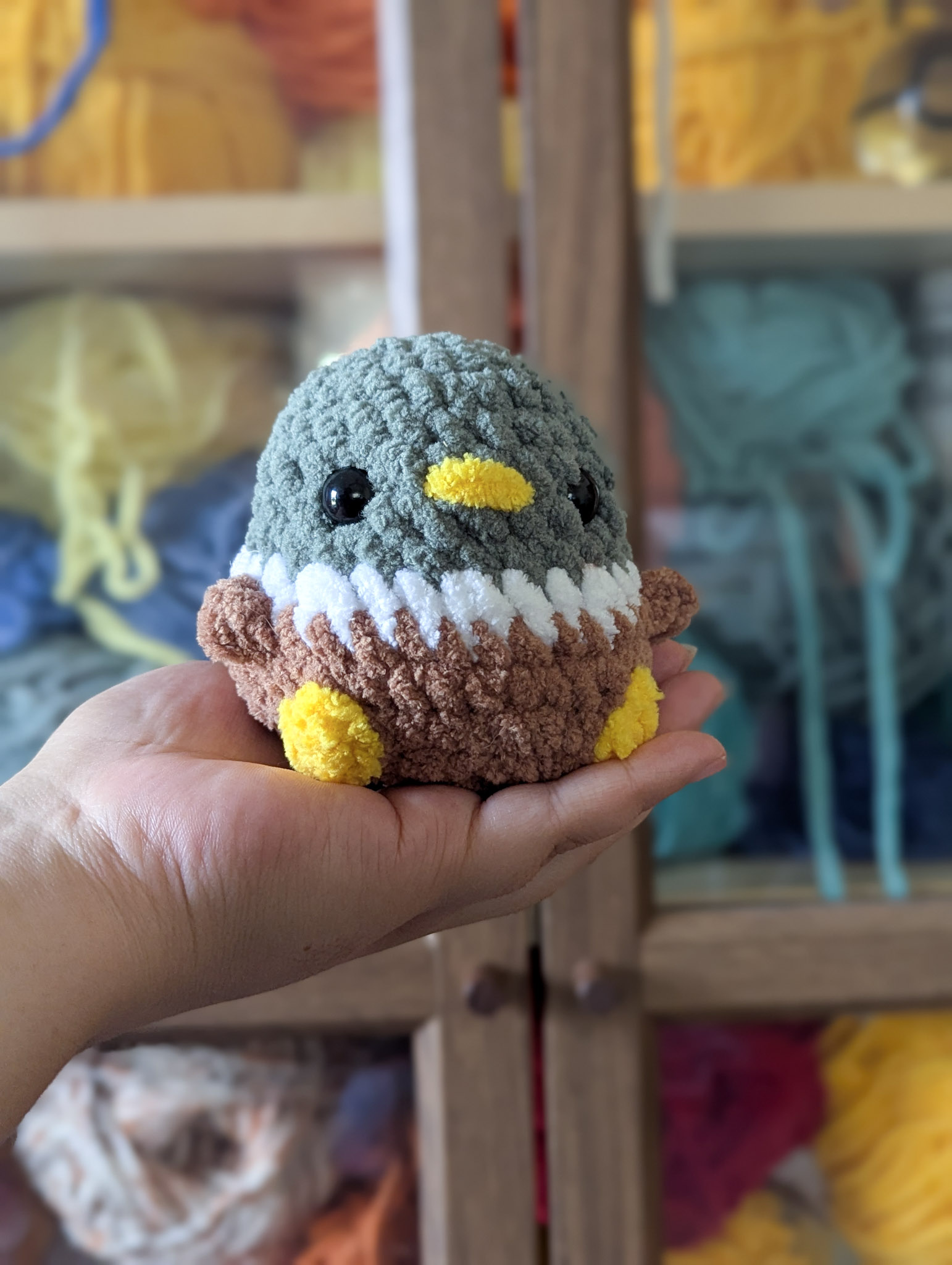 Small classic chubby duck plushie being held in front of a backdrop of yarn in a cabinet.