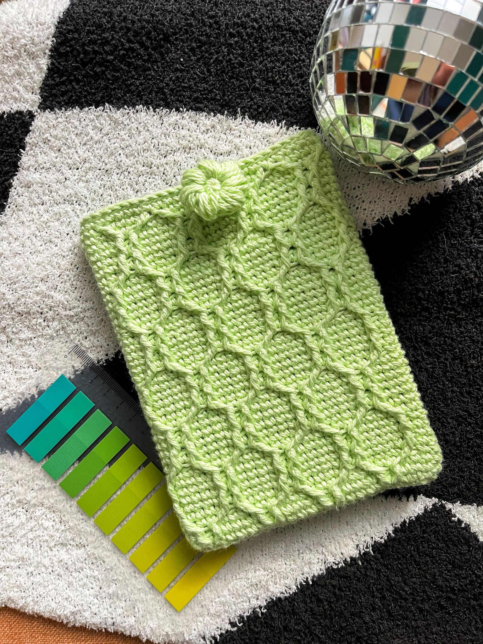 a bright, lime green crocheted Kindle sleeve with a diamond ribbing texture. Next to it lays a set of sticky tabs and a small shiny disco ball for an aesthetic look.