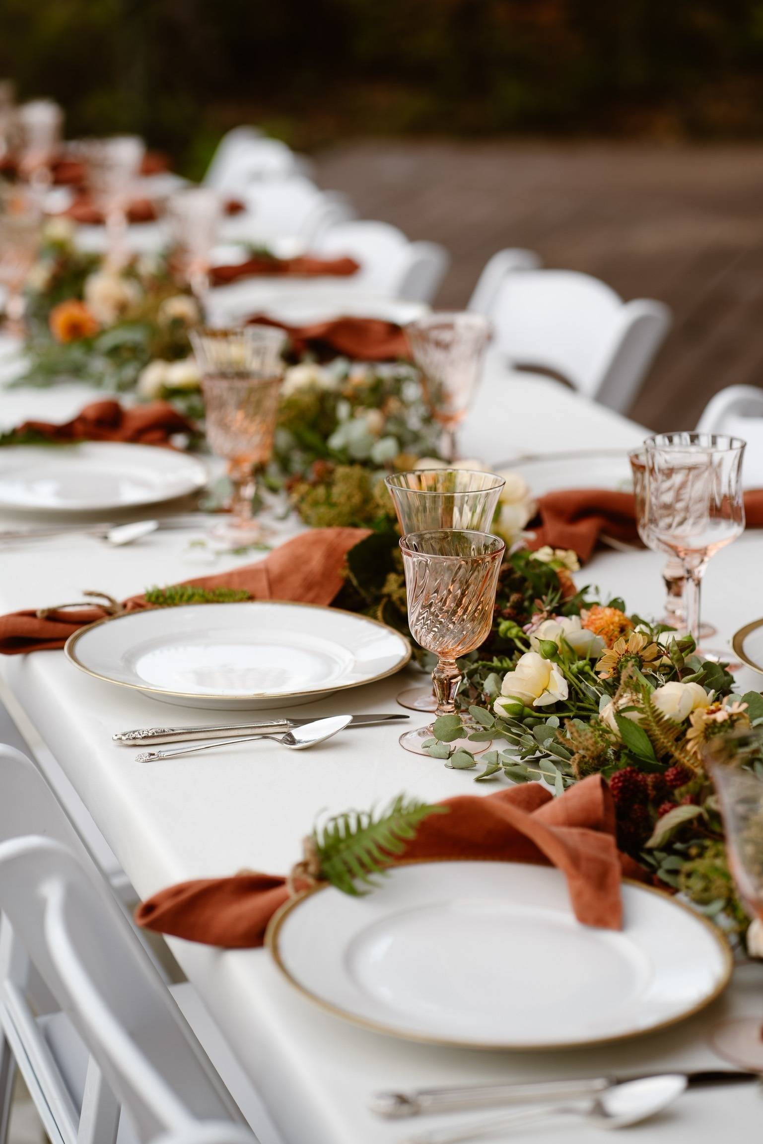 Table set with white plates with gold rims, blush pink glassware, cinnamon colored linen napkins tied with twine and a tiny fern with a table runner of fresh florals down the center.