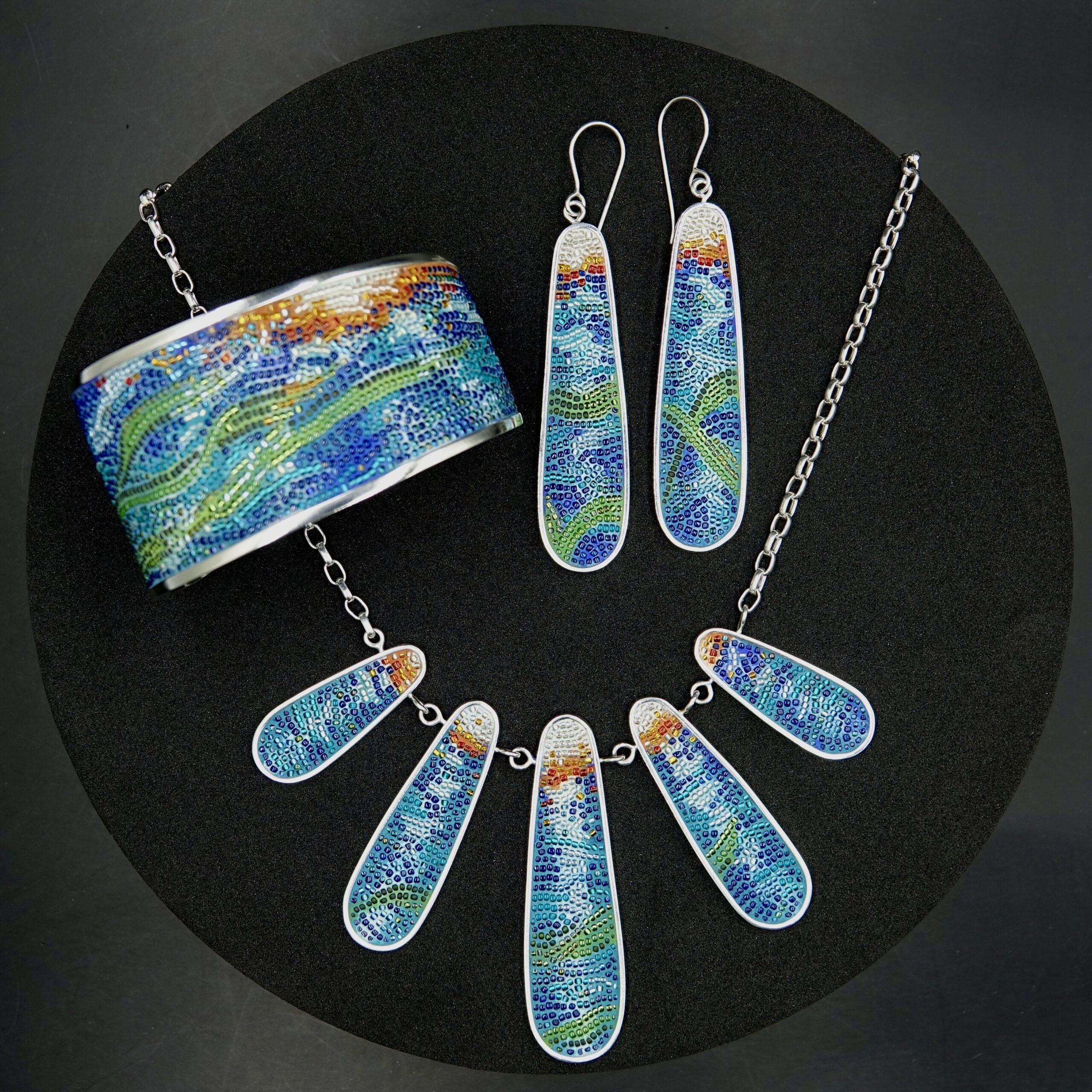 A micro-mosaic jewelry suite including necklace, earrings and bracelet, with abstract imagery of sunshine, waves and kelp. The necklace has five long teardrop shaped graduated pieces hanging from a chain, the earrings echo that shape, in the largest size, and the bracelet is a wide cuff style.