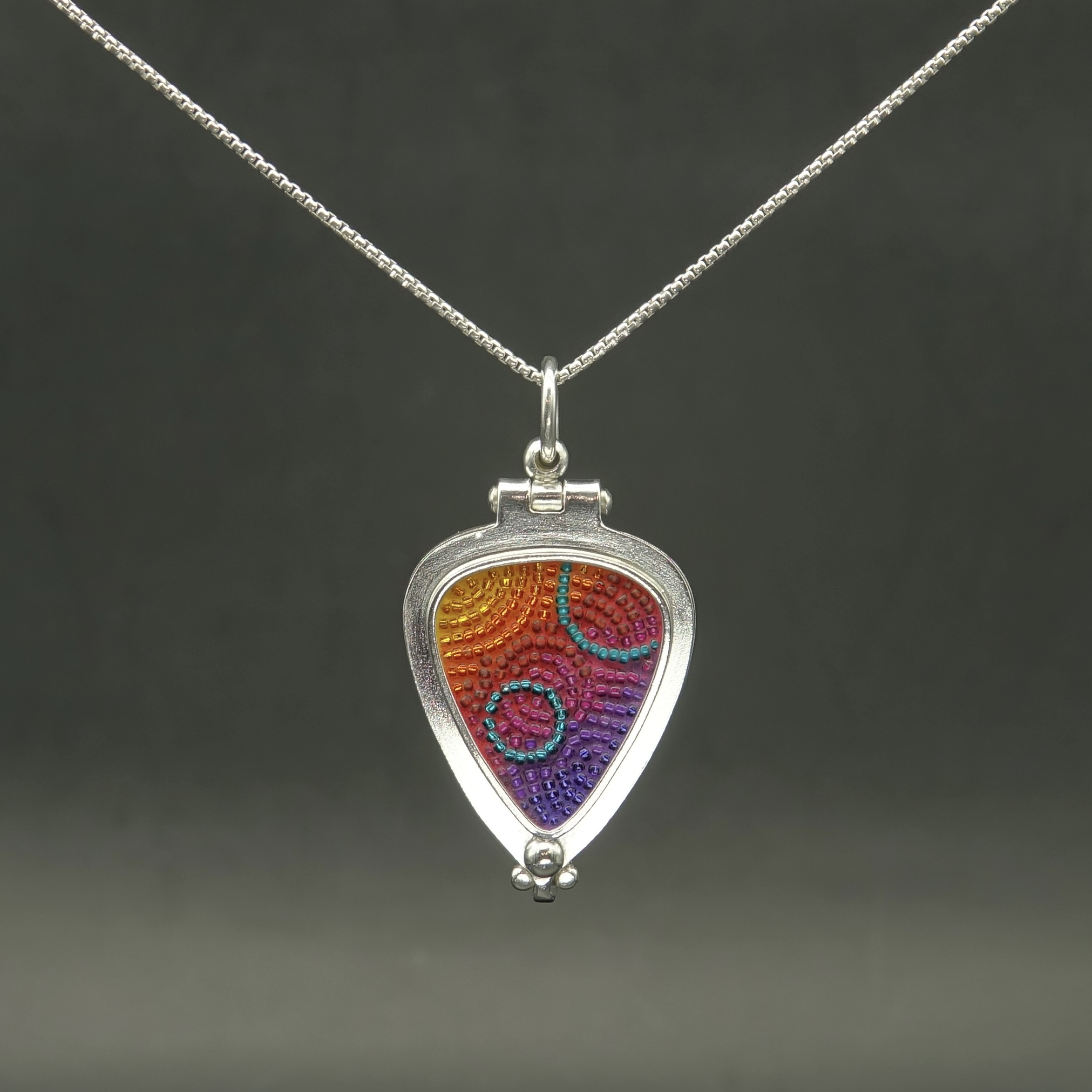 A guitar pick-shaped locket with colorful micro-mosaic inlay made of sterling silver and glass beads, ranging from blues, purples, reds and oranges. There is a hinge at the top and a clasp at the bottom.