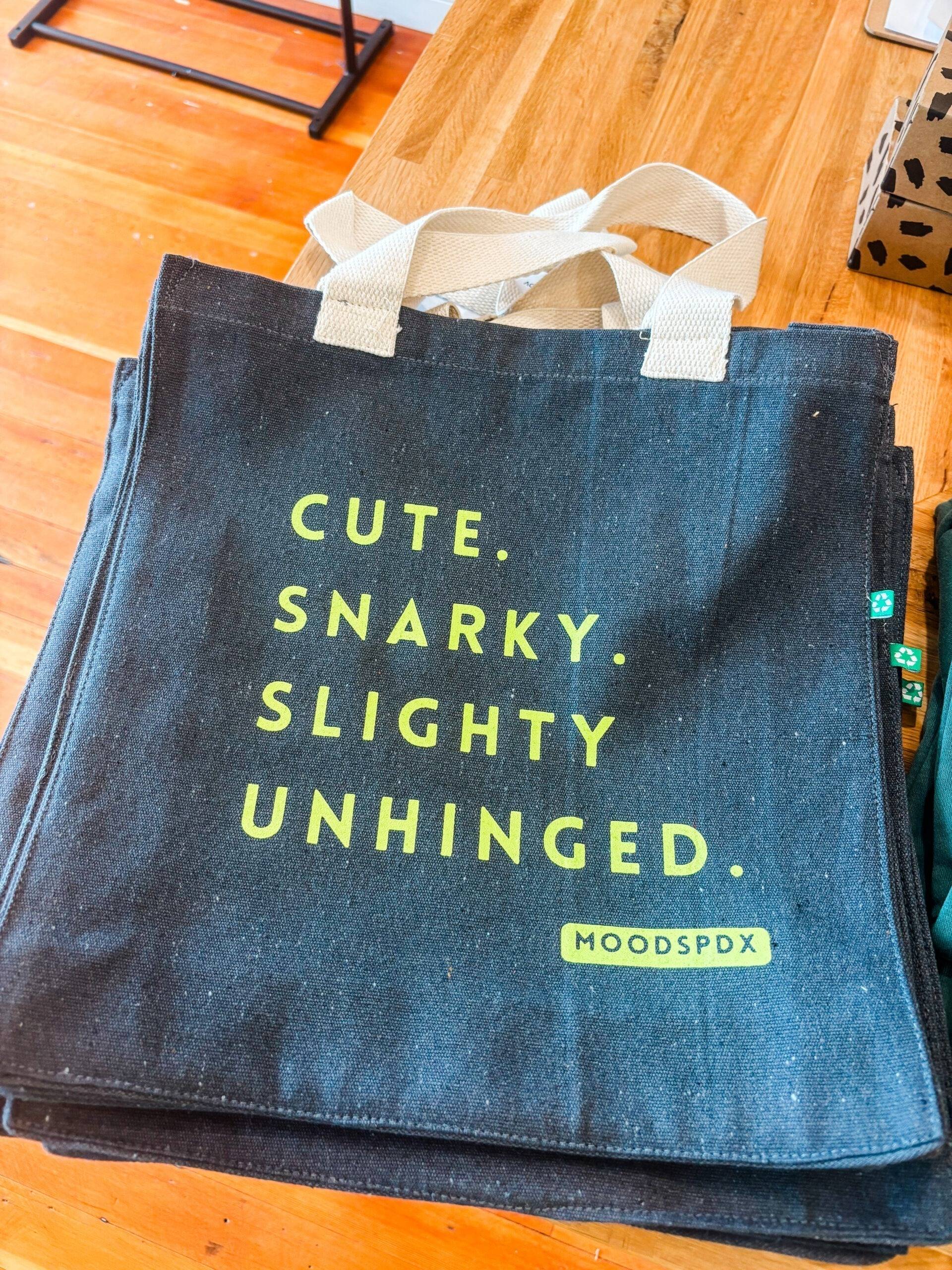 Grey canvas shopping tote bag screen printed with the phrase “cute, snarky, slightly unhinged.”
