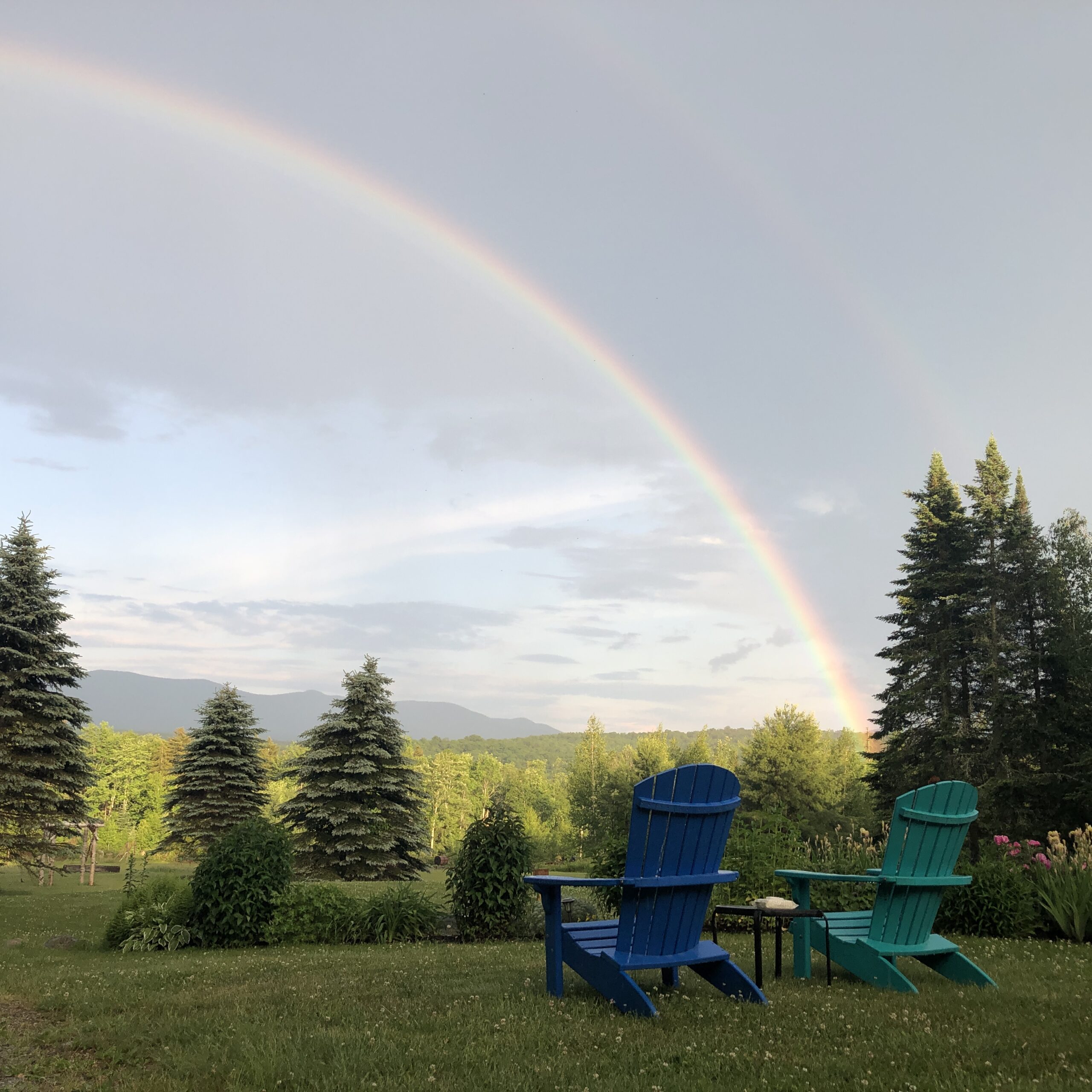 Double rainbow above two Adirondack chairs placed in the meadow.