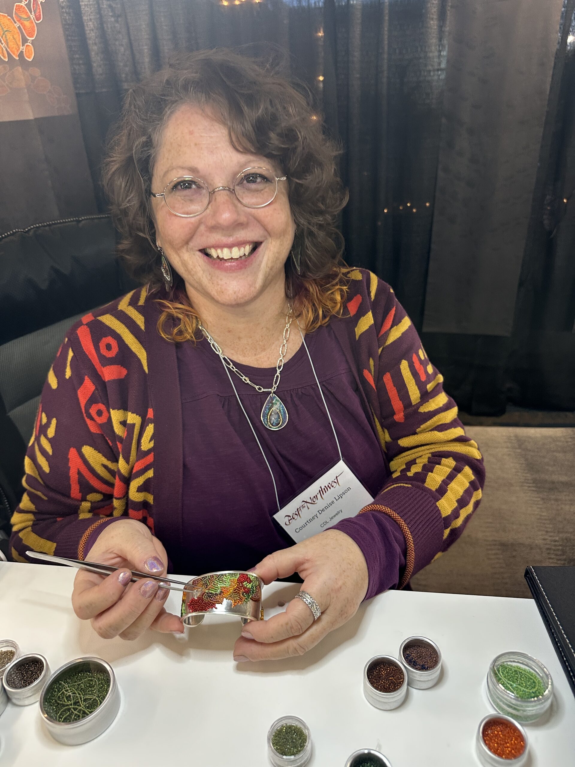 A woman wearing a plum sweater with mustard yellow and orange patterns, sits with her hands holding a bracelet she is inlaying with tiny glass beads using a pair of tweezers.