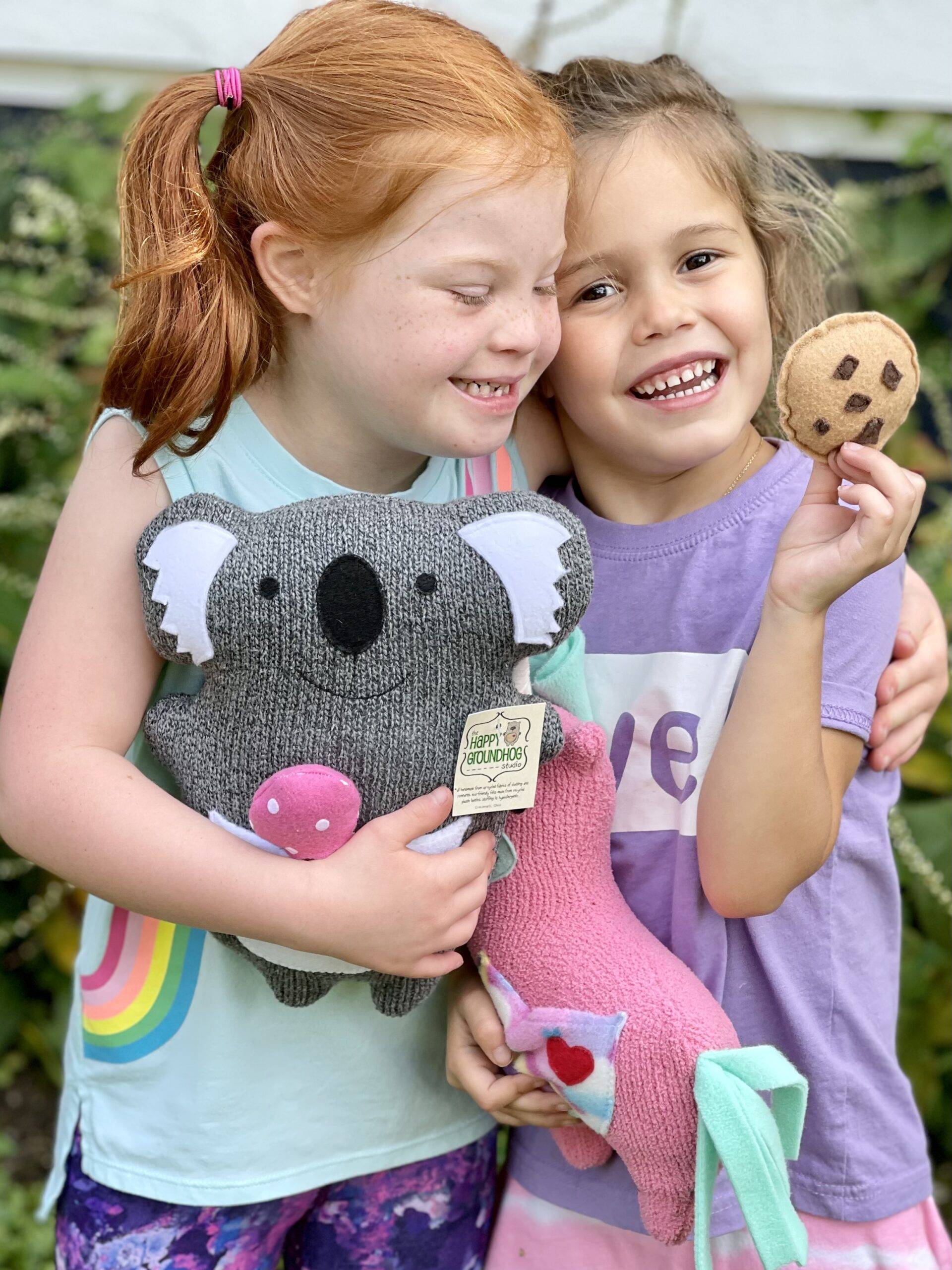 Two little girls smiling. one with brown hair the other has red hair and Down syndrome both holding cute handmade stuffed animals