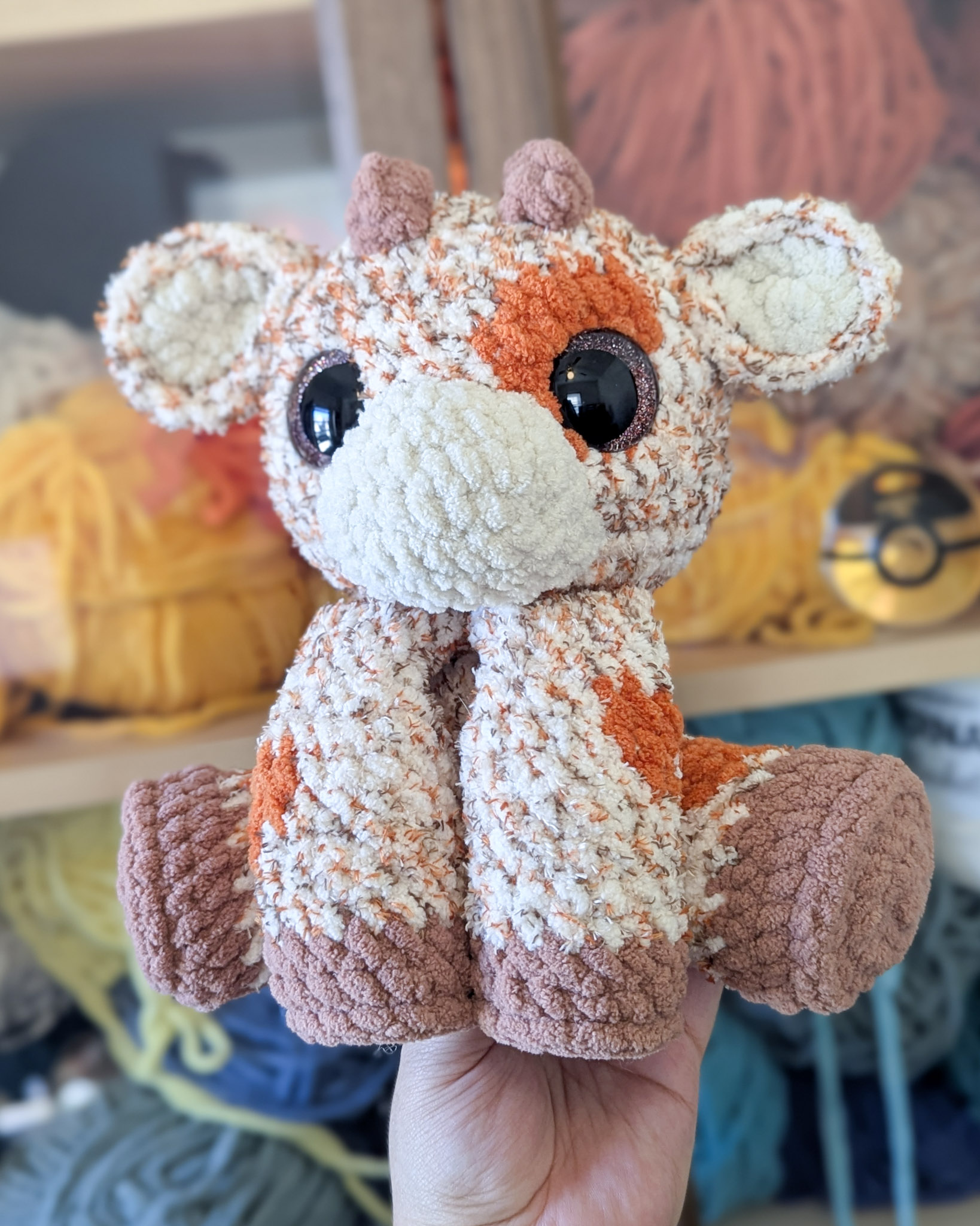 Large pumpkin spice latte inspired cow plushie being held in front of a backdrop of yarn in a cabinet.