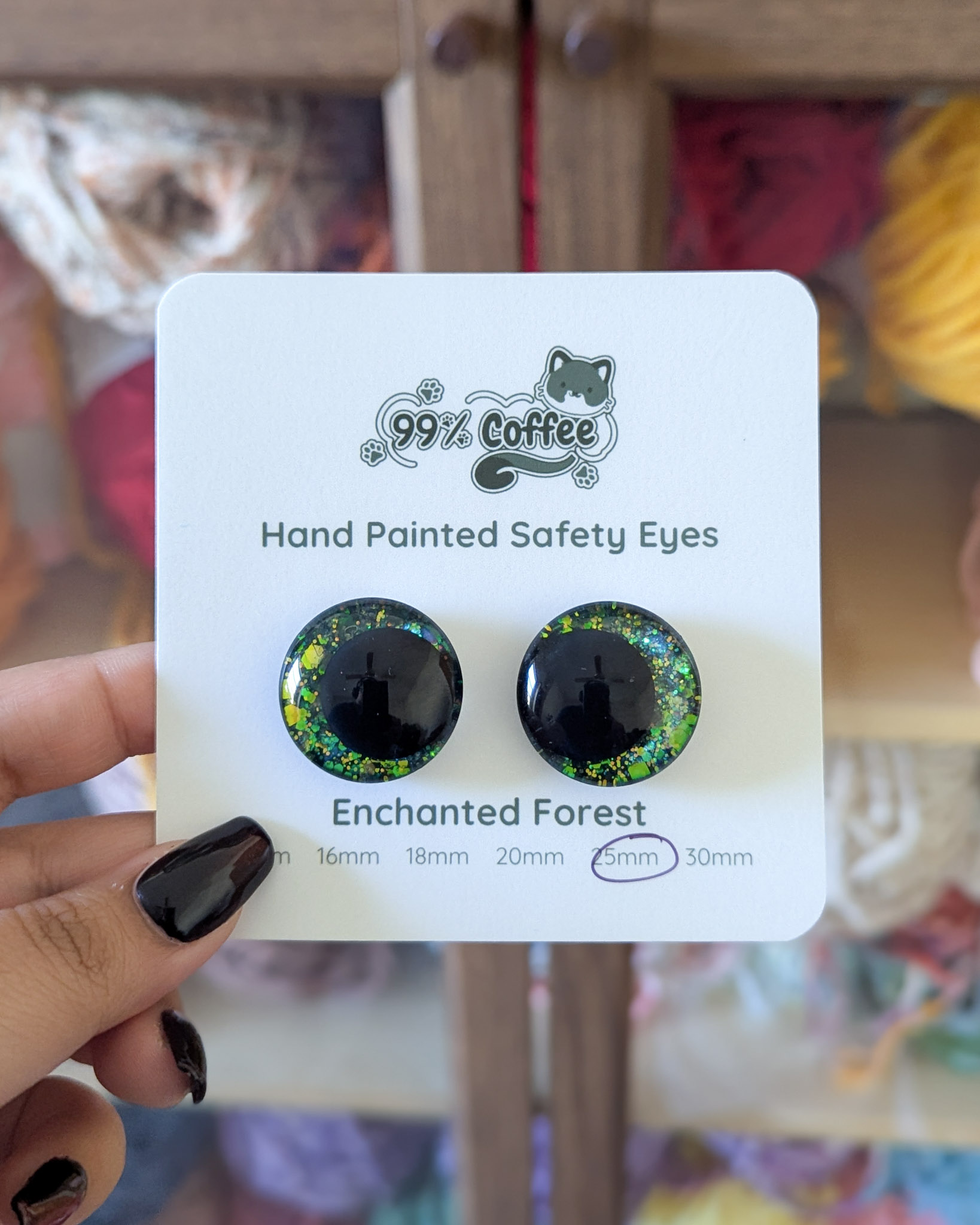 A pair of green glitter safety eyes on a white card being held in front of a backdrop of yarn in a cabinet.