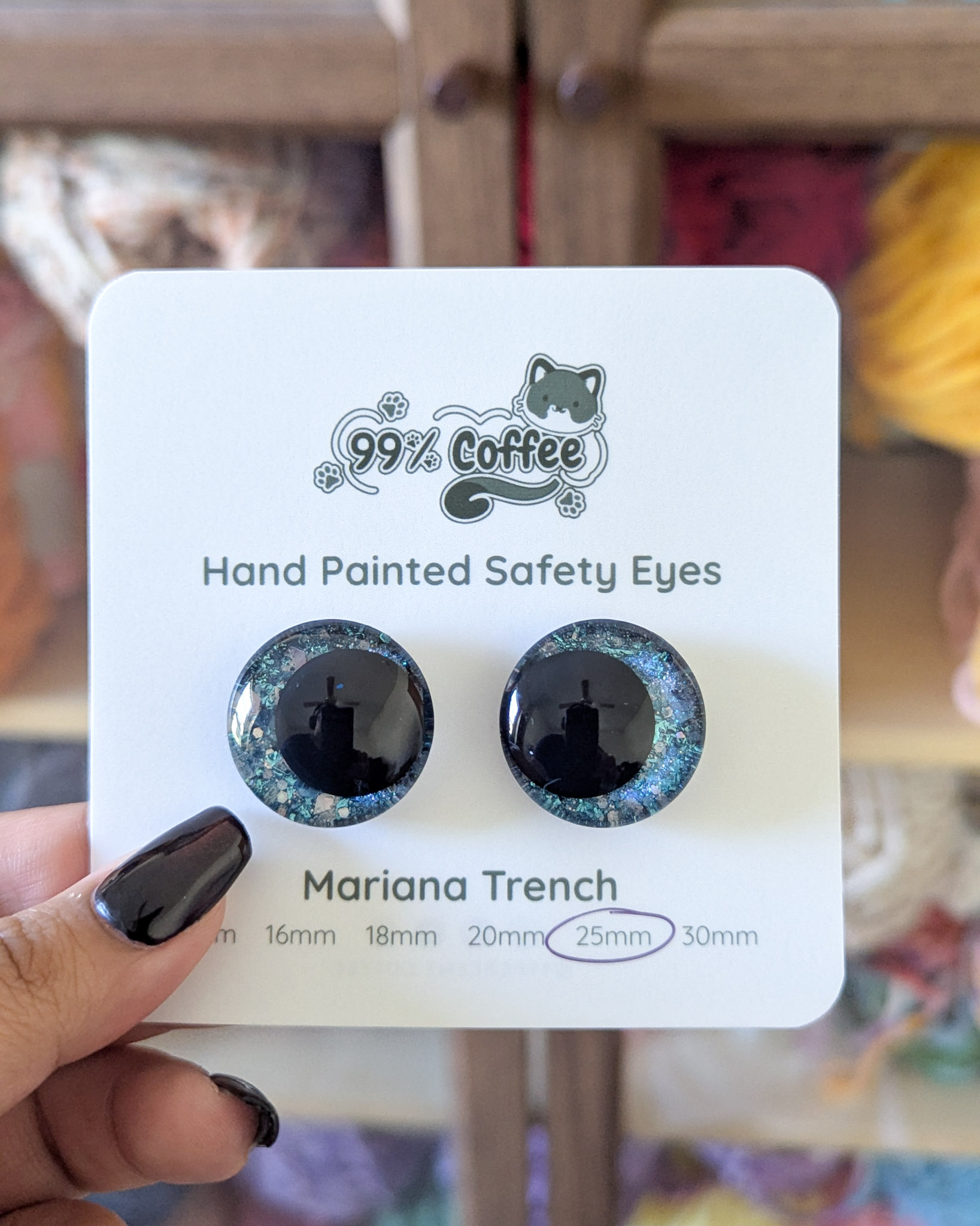 A pair of blue glitter safety eyes on a white card being held in front of a backdrop of yarn in a cabinet.