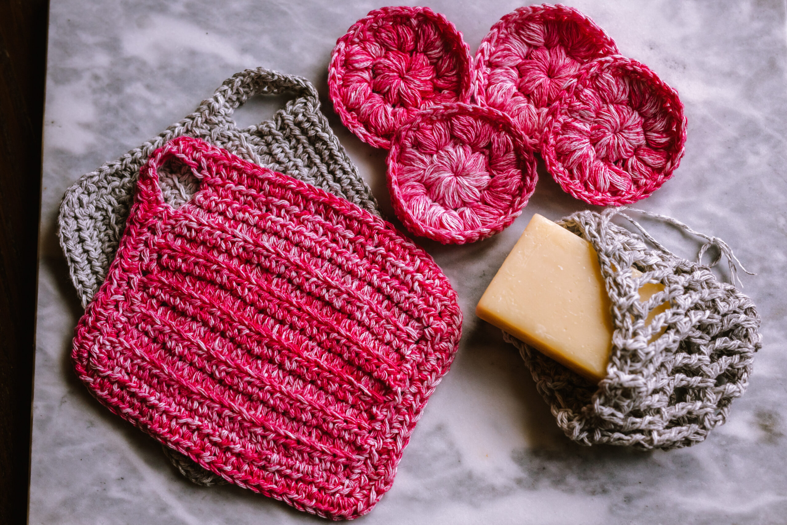 An image of two handmade washcloths, four facial scrubbies, and a soap saver with a bar of soap in it. The items are a mix of pink yarn and grey yarn.