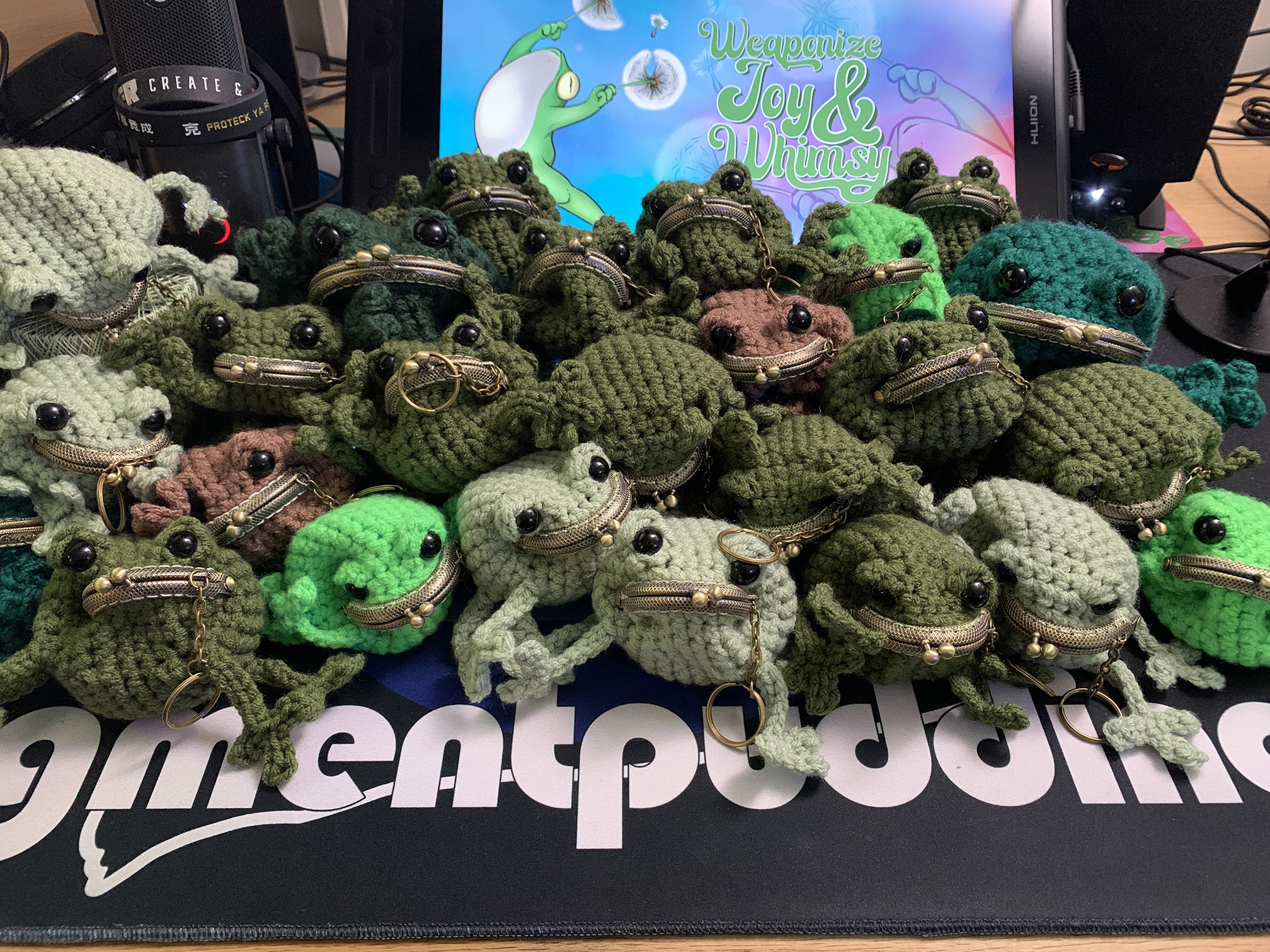 A pile of small crocheted frog coin purses in various shades of green, sitting in front of a digital tablet screen with a brightly colored image of a leaping frog, and text that says