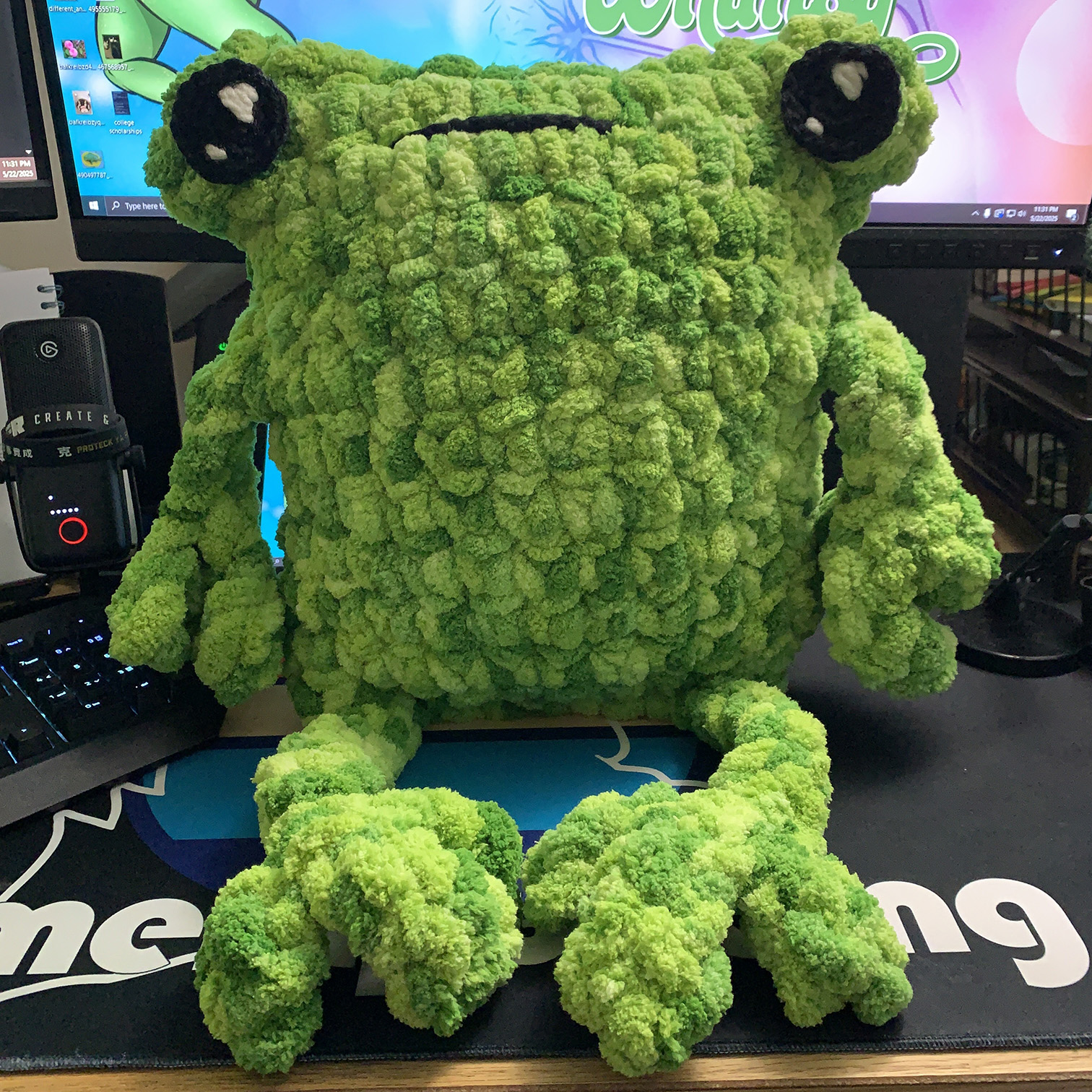 A large bright green throw pillow frog sitting on a desk. His body is very square, and he has big squishy hands and feet.
