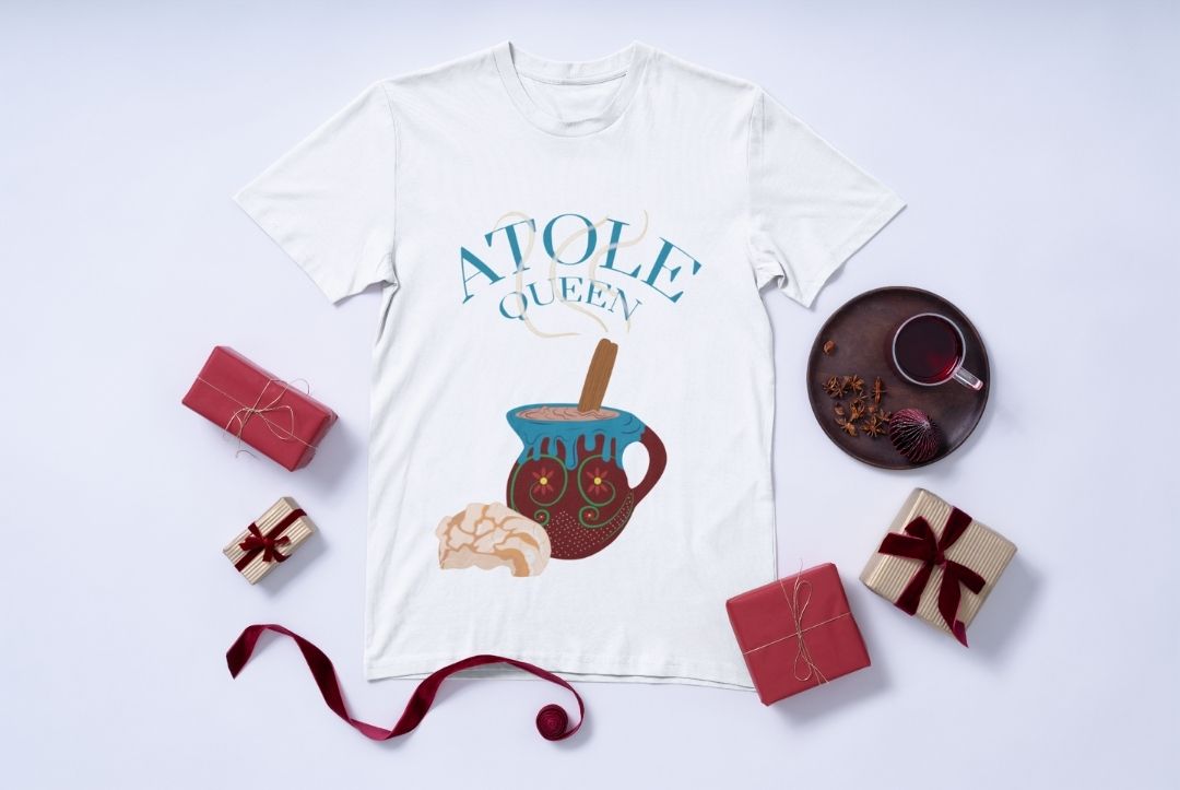 White t-shirt with a colorful illustration of a clay mug of atole, a cinnamon stick, and a concha, with the text ‘Atole Queen’ above it.