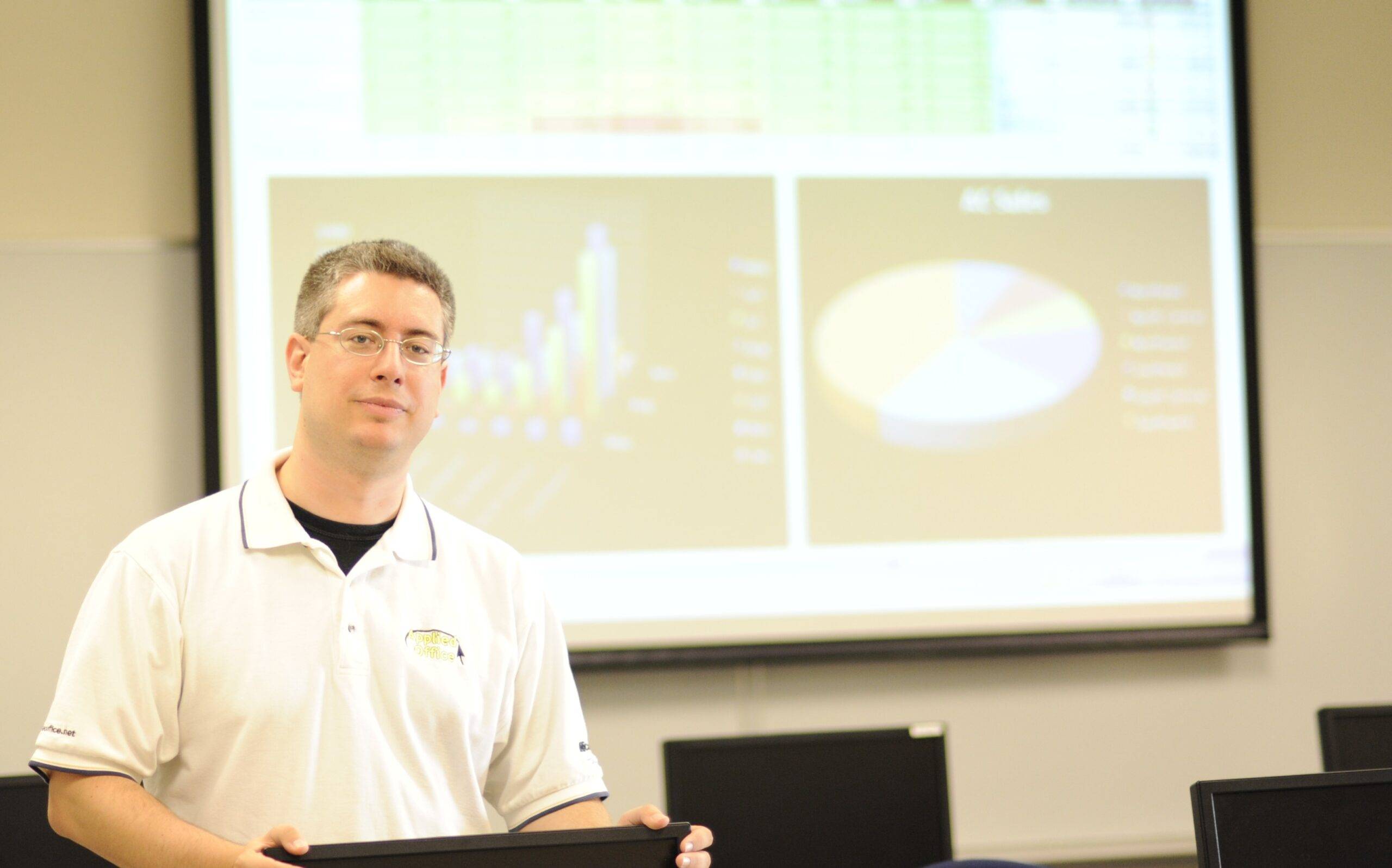 David, the owner, is in a classroom in front of a projection screen showing Microsoft Excel during a class.