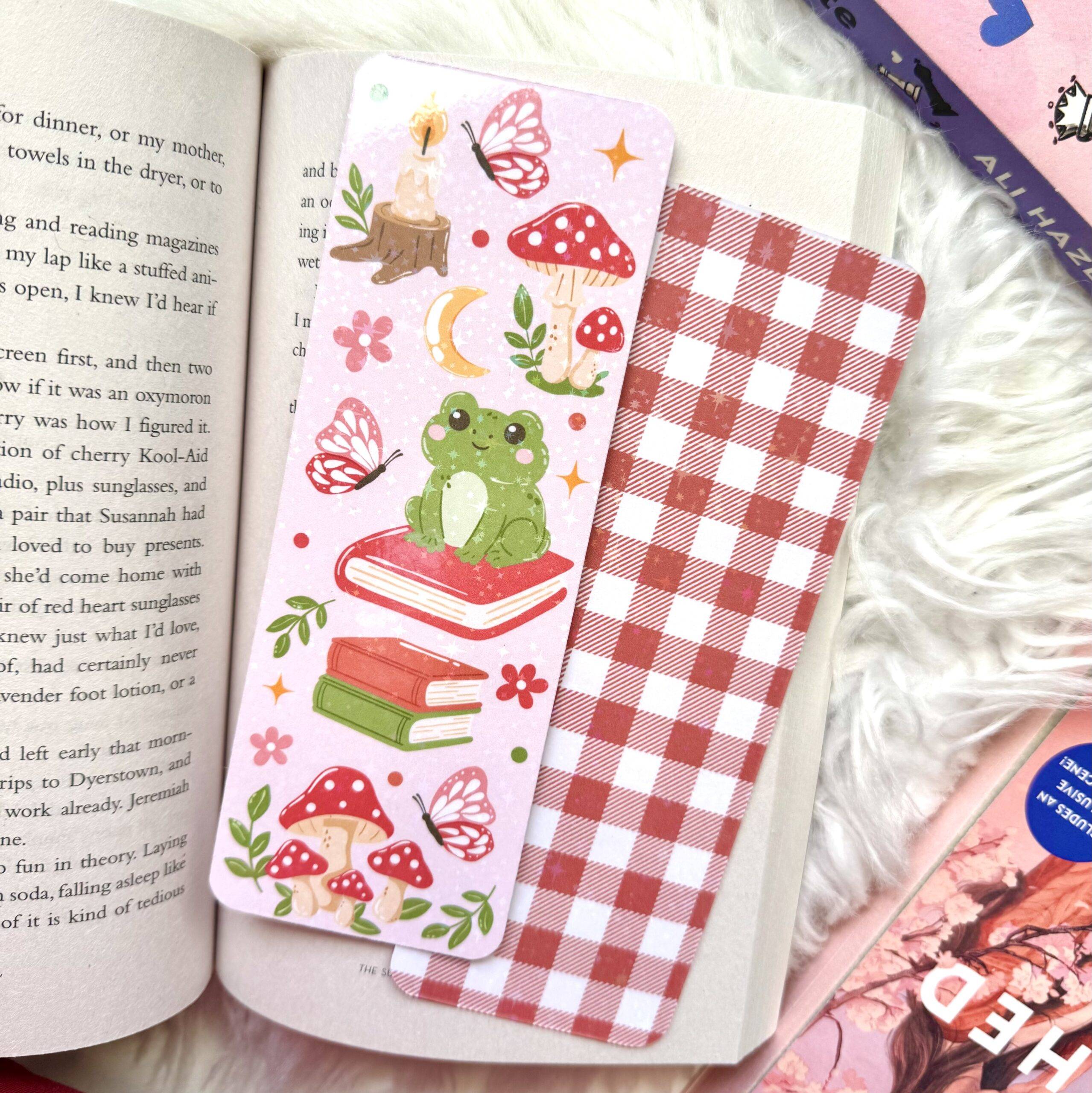 A pink bookmark illustrated with a frog, mushrooms, and bookish elements resting upon an open book.