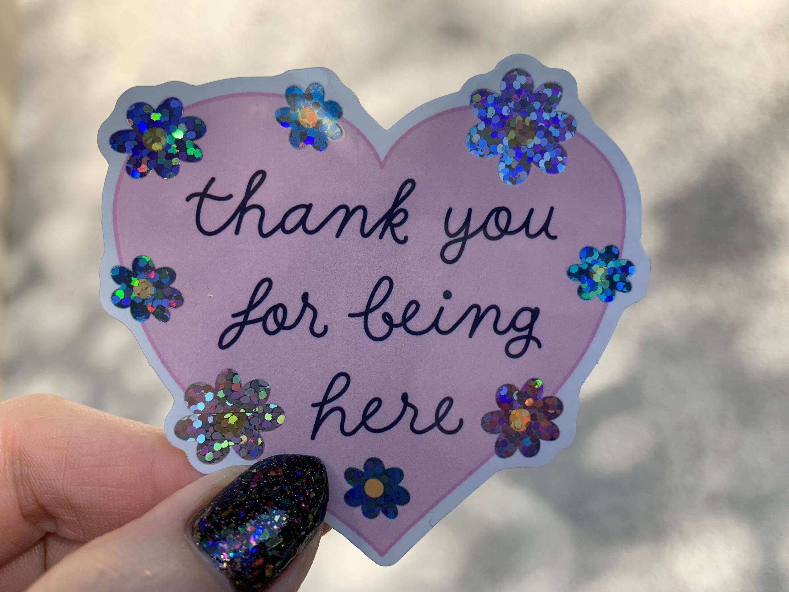 Glossy pink heart sticker with blue script reading, “thank you for being here.” Script is surrounded by glittery flowers.