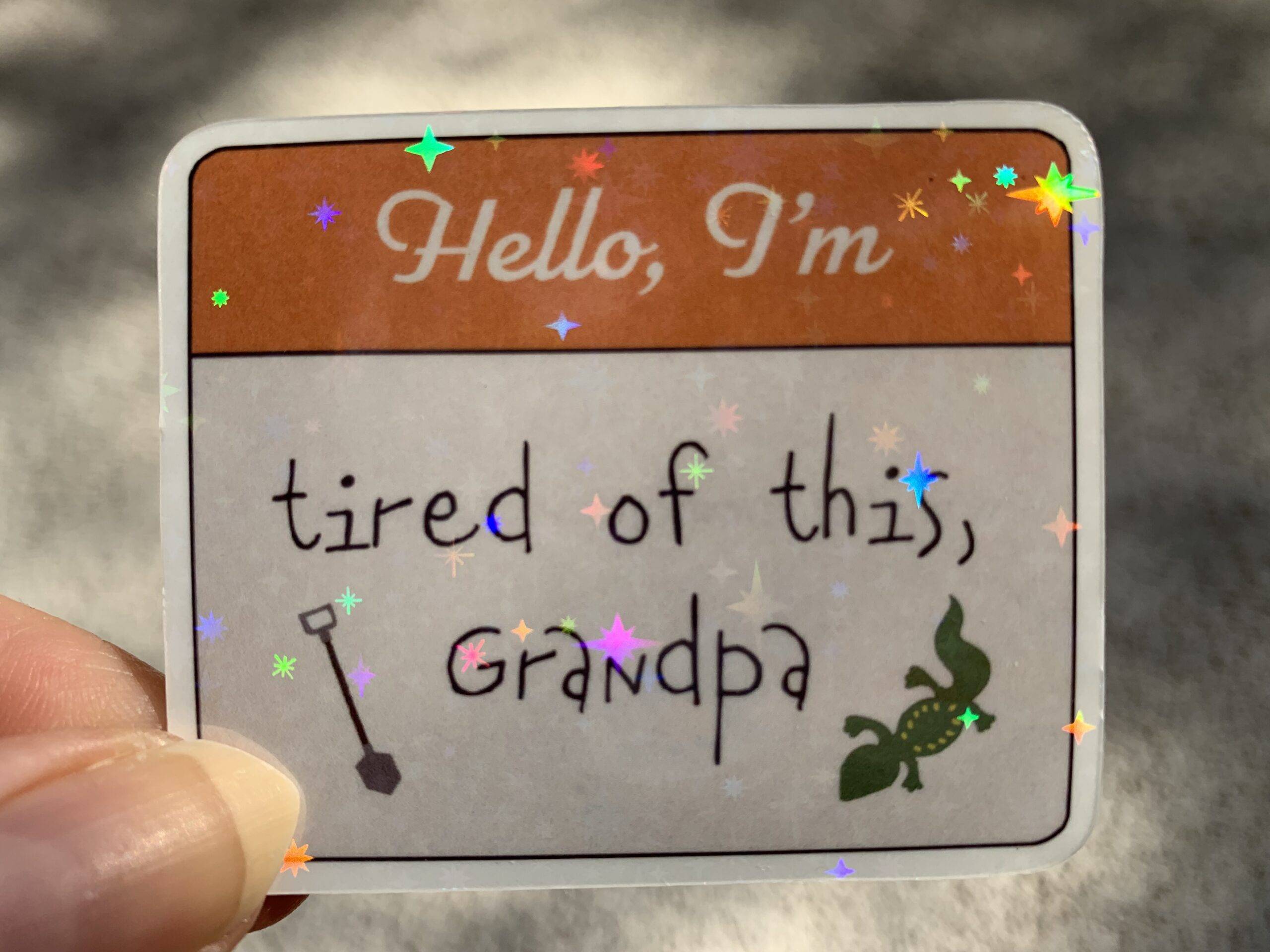 A hand holding a sticker with twinkle glitter finish reading, “hello, I’m tired of this, Grandpa” with illustrations of a shovel and a lizard.