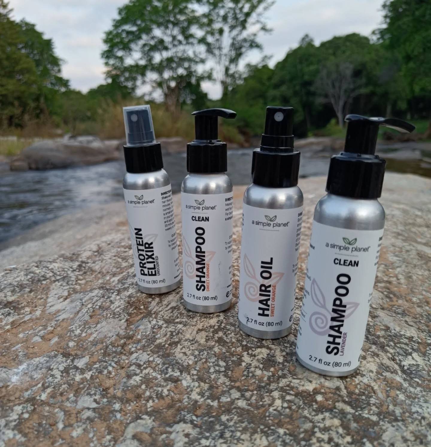 Several A Simple Planet aluminum hair-care bottles sitting on a natural rock beside a creek, photographed outdoors with trees in the background.