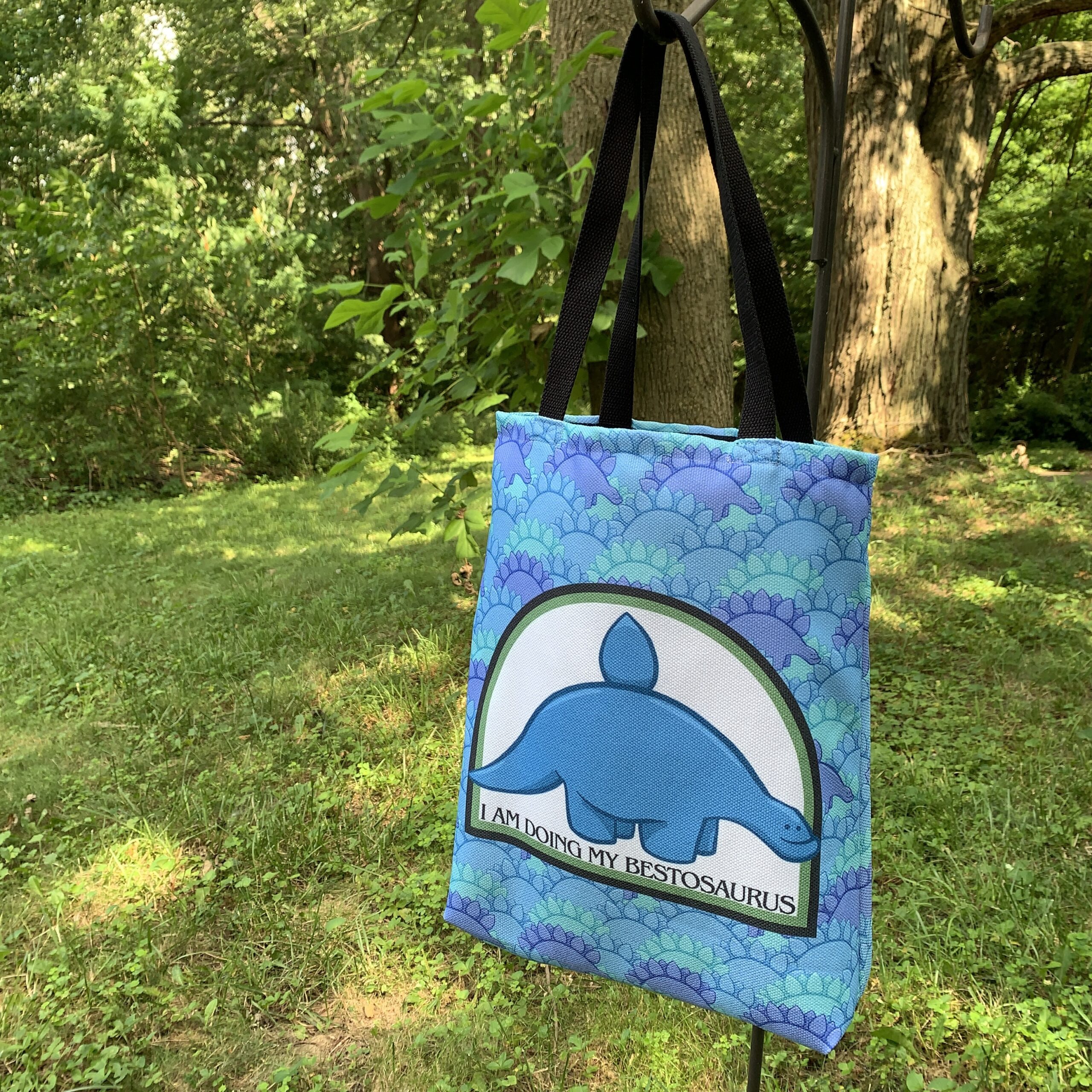 A tote bag hangs in front of a woodsy background. A pattern of stegosauruses cover the bag, with a central image of a blue stegosaurus with a single scute and a bit of an underbite. Text beneath says