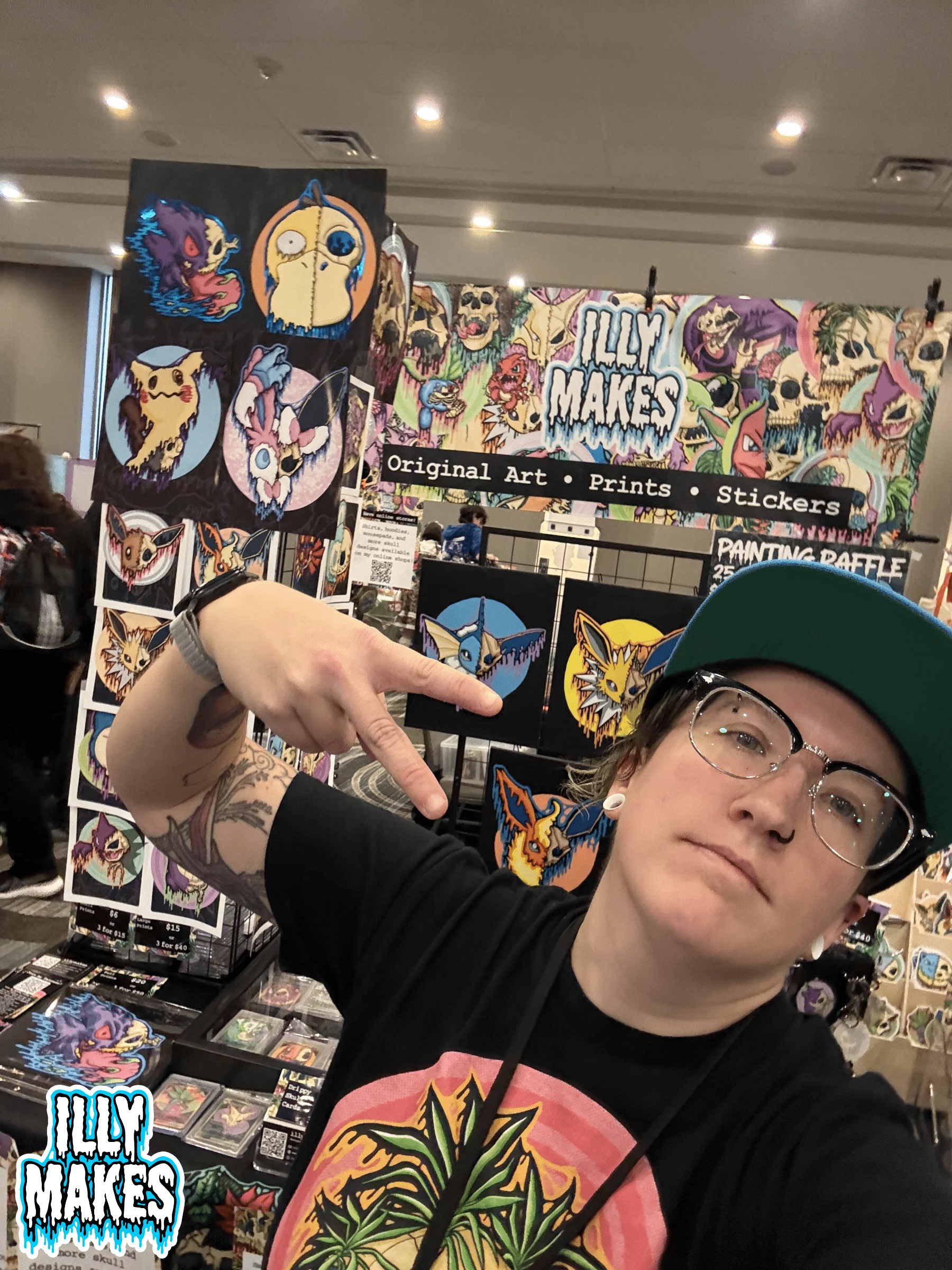Me standing in front of the Illymakes convention booth, surrounded by drippy skull prints and colorful artwork, posing with a peace sign.