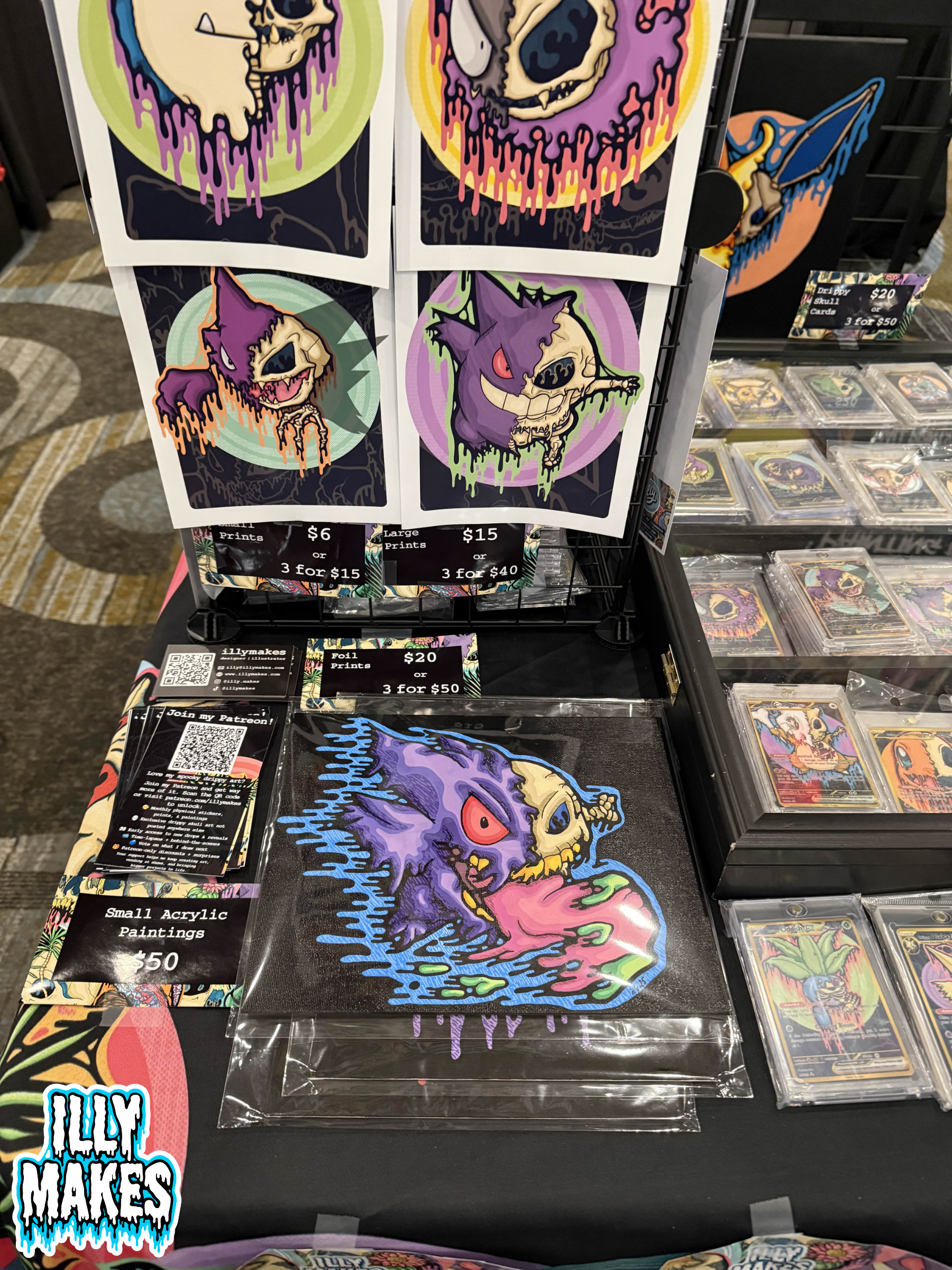 Illymakes convention table display featuring drippy skull Pokémon prints, sticker packs, small acrylic paintings, and a pricing sign arranged on a black table.
