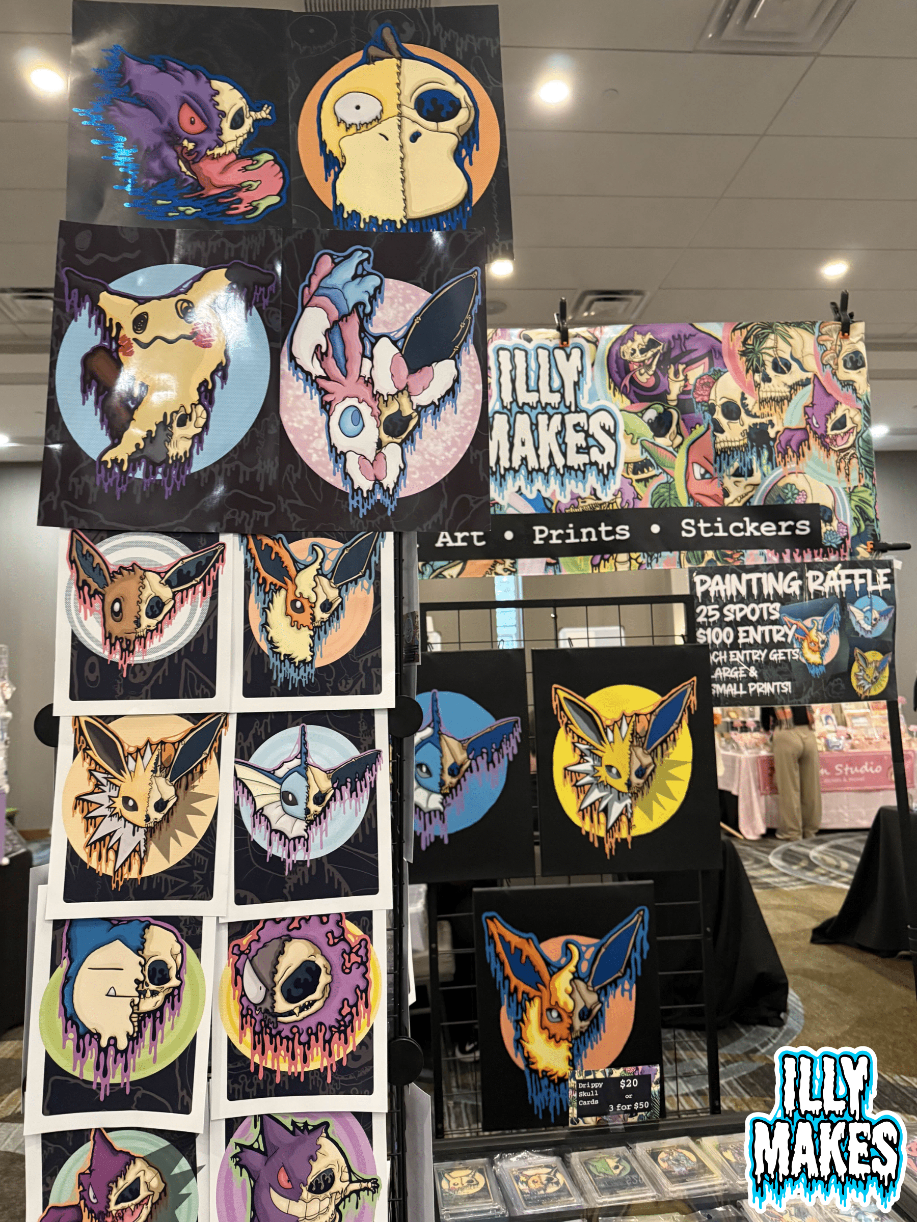 Illymakes booth display with large drippy Pokémon skull prints arranged on a grid wall, plus a colorful illustrated banner featuring skull-themed artwork.