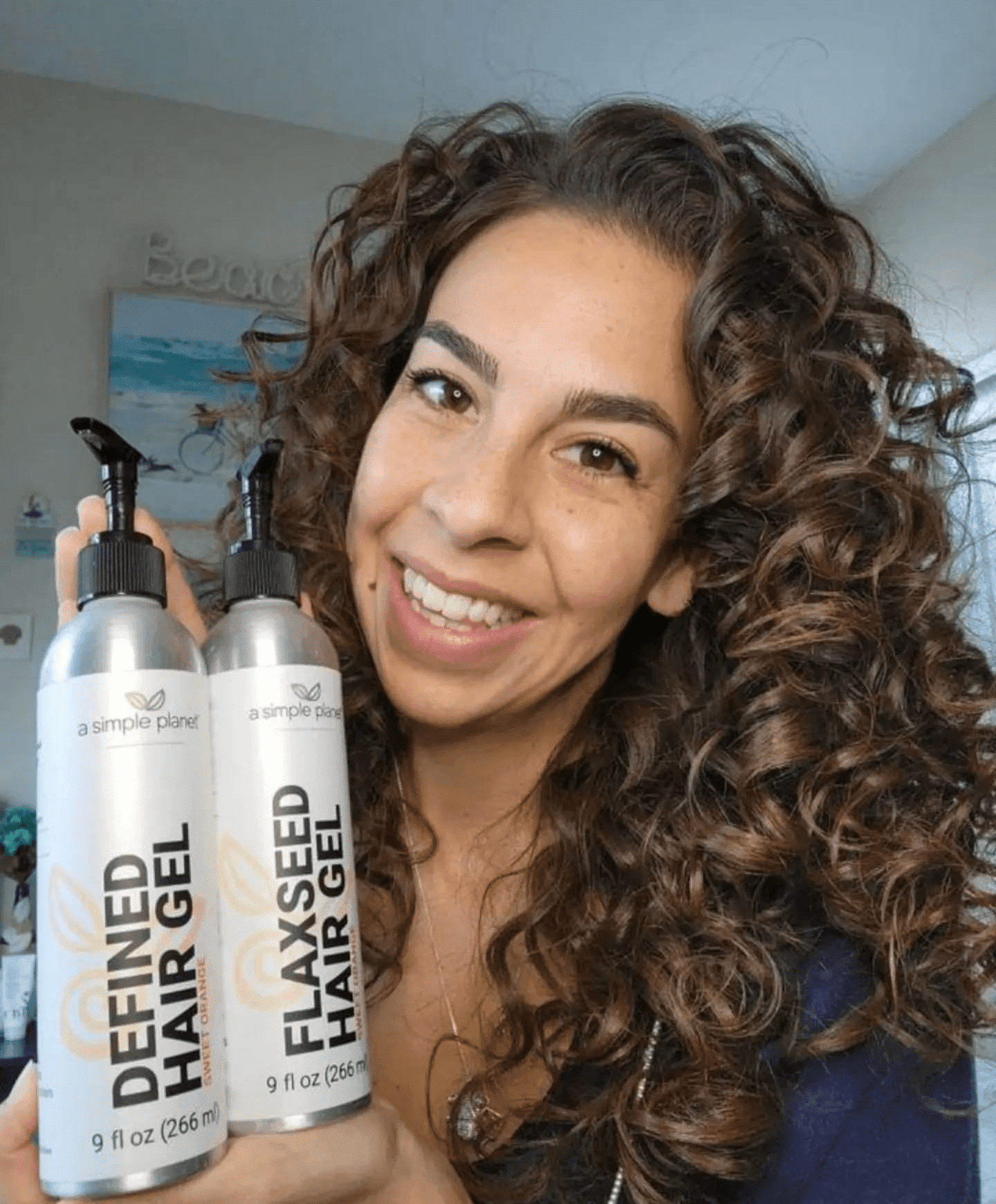 A smiling woman with long, defined curls holds two A Simple Planet aluminum hair gel bottles in front of her.