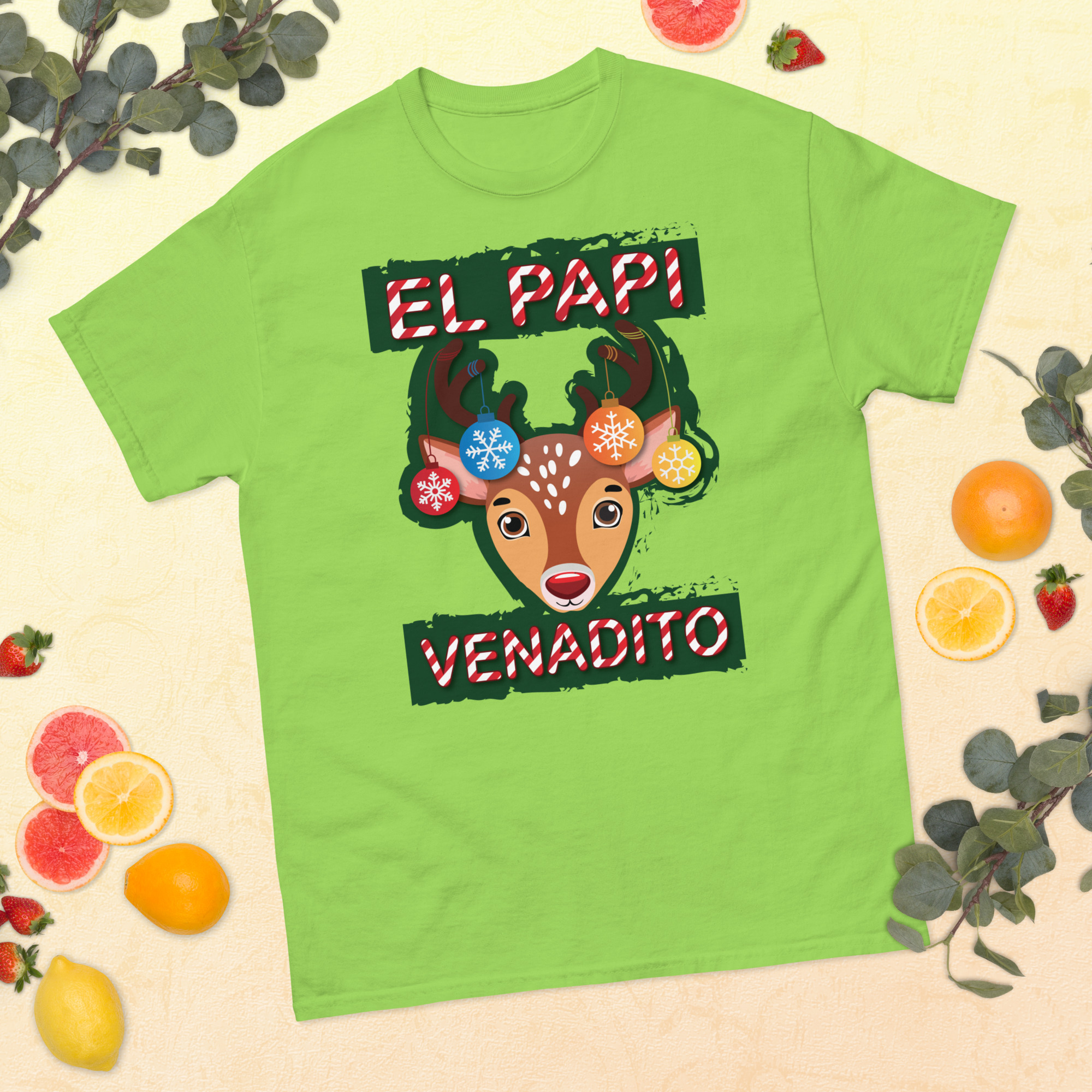 Flat lay of a lime green t-shirt featuring a cute reindeer illustration with holiday ornaments on its antlers and the phrase ‘El Papi Venadito’ in candy cane lettering.