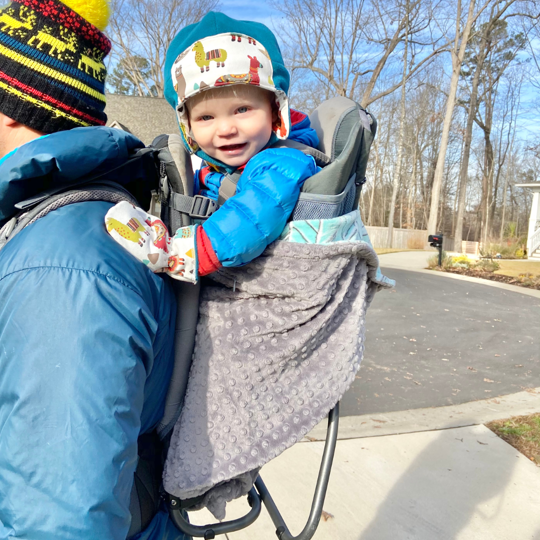 A happy, young child in a hiking backpack, bundled up on a cold winter day, with a grey Cocoona around the outside of the pack keeping kiddo warm.