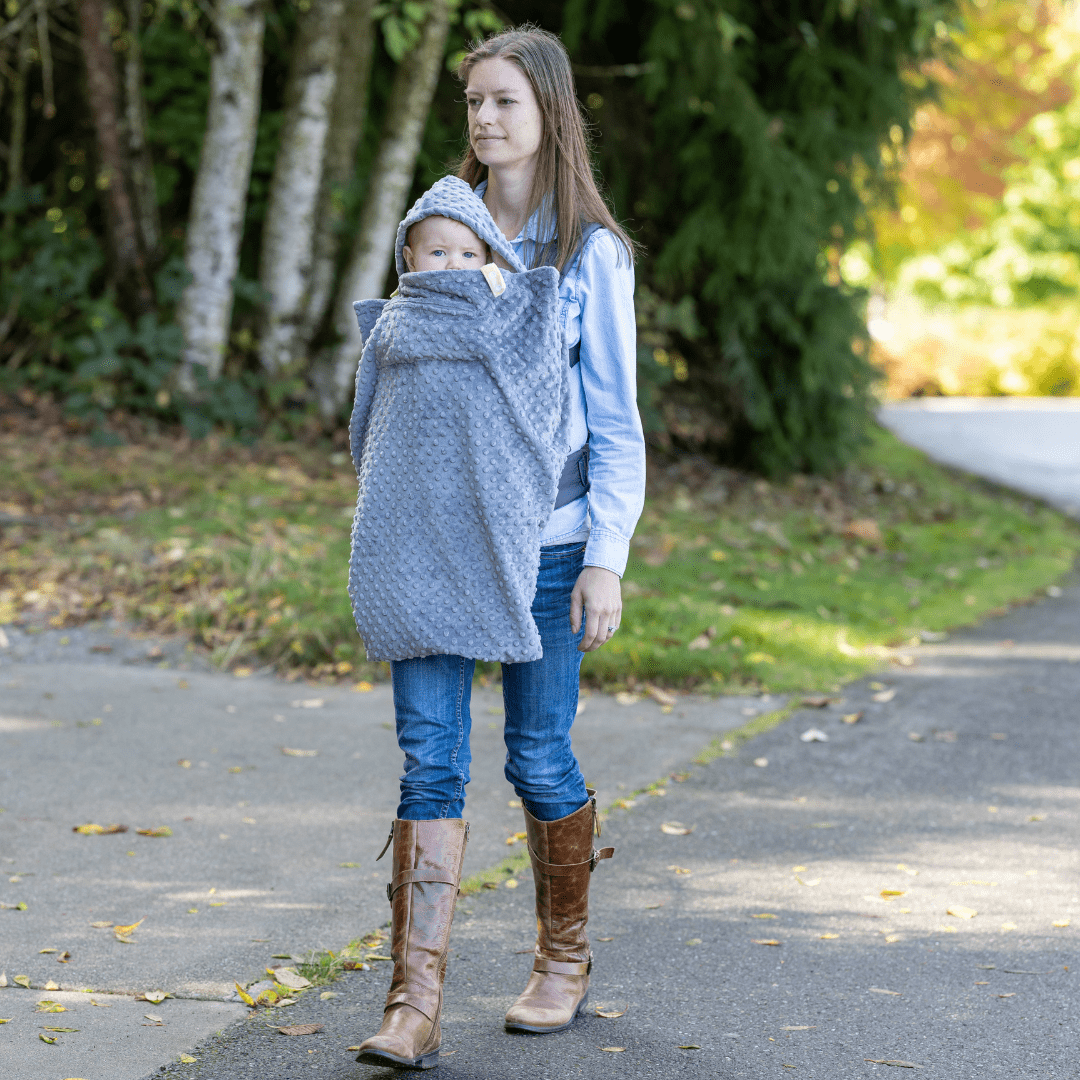 A woman walking through the park on a fall day wearing her baby in a carrier on her chest. The baby has a Cocoona enveloping their body and a hood on their head for warmth.