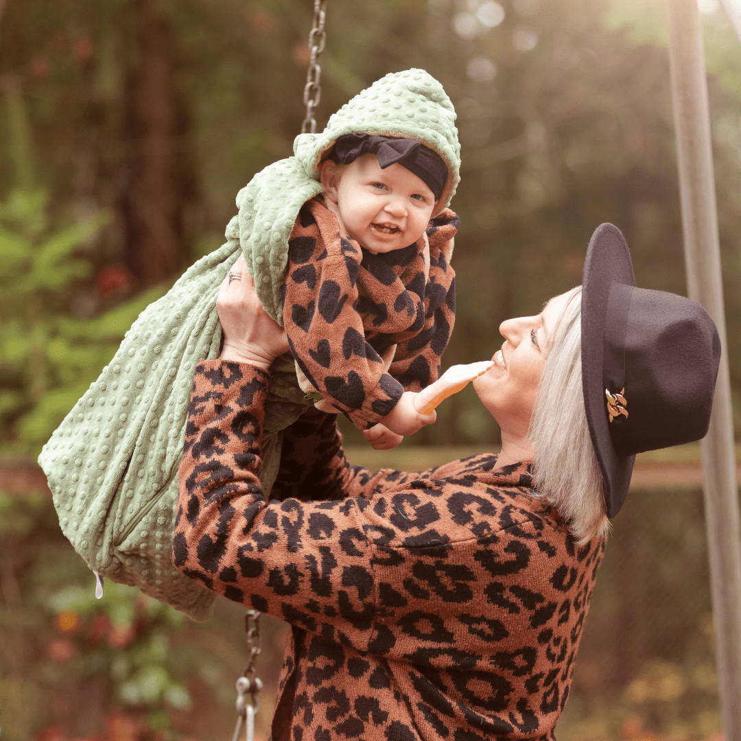 A fall day playing at the park, a mom holding up her smiling baby whose body is enveloped in a Cocoona with the hood on.