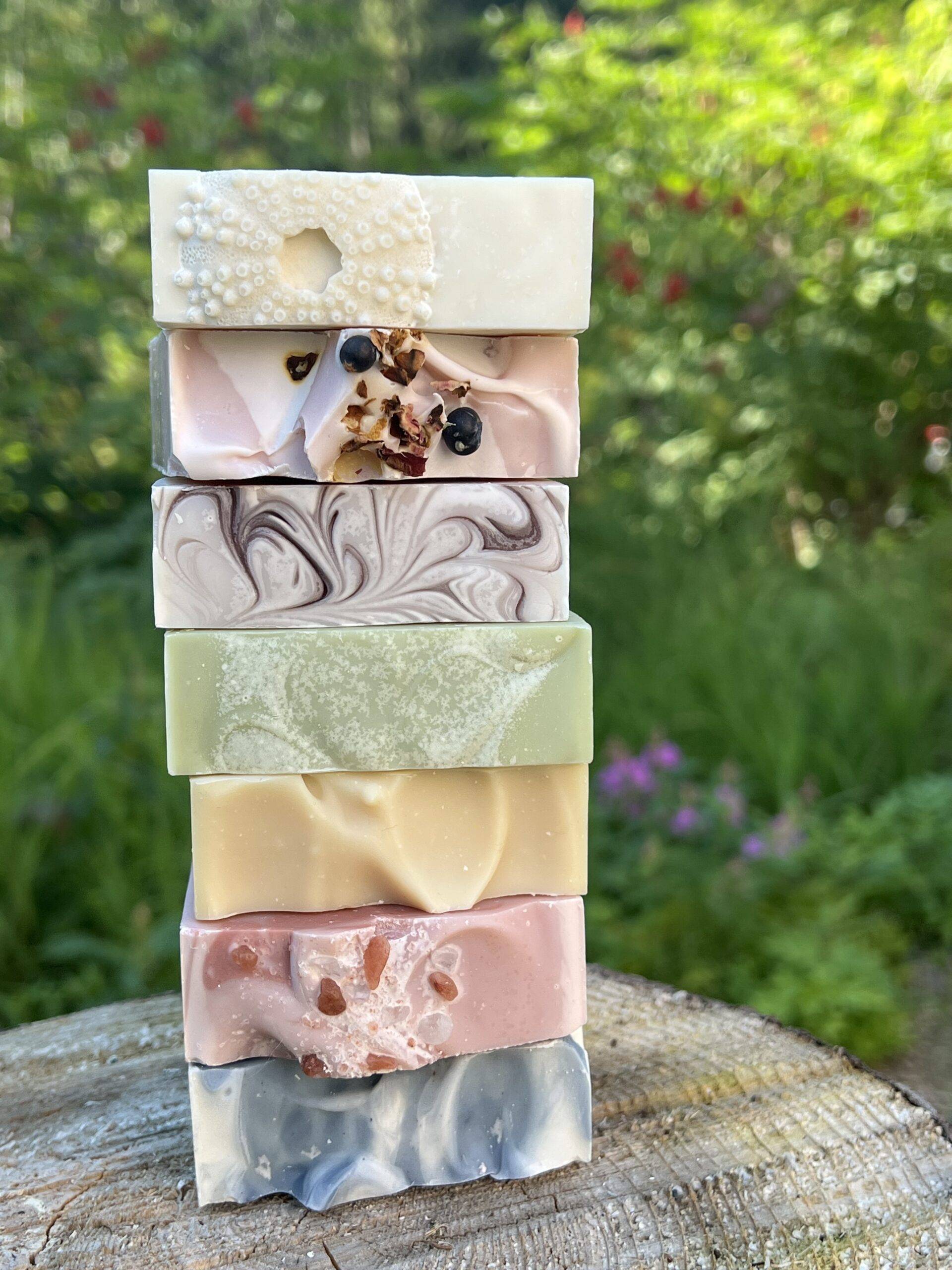 Seven different soap bars stacked on the cut end of a log. Wild berry bushes and fireweed are visible in the background.