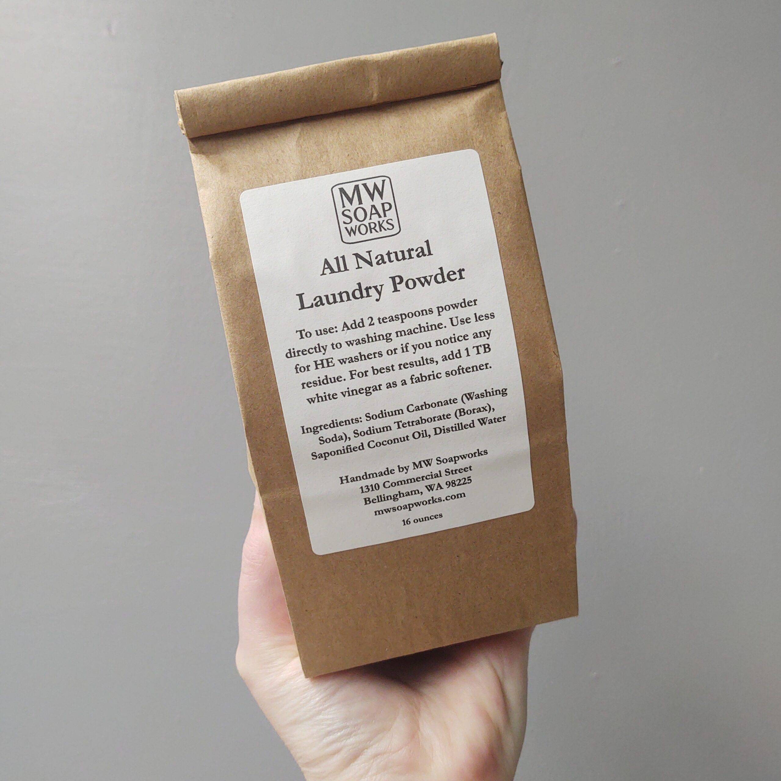 A hand holds up a brown paper bag of MW Soapworks All Natural Laundry Powder.