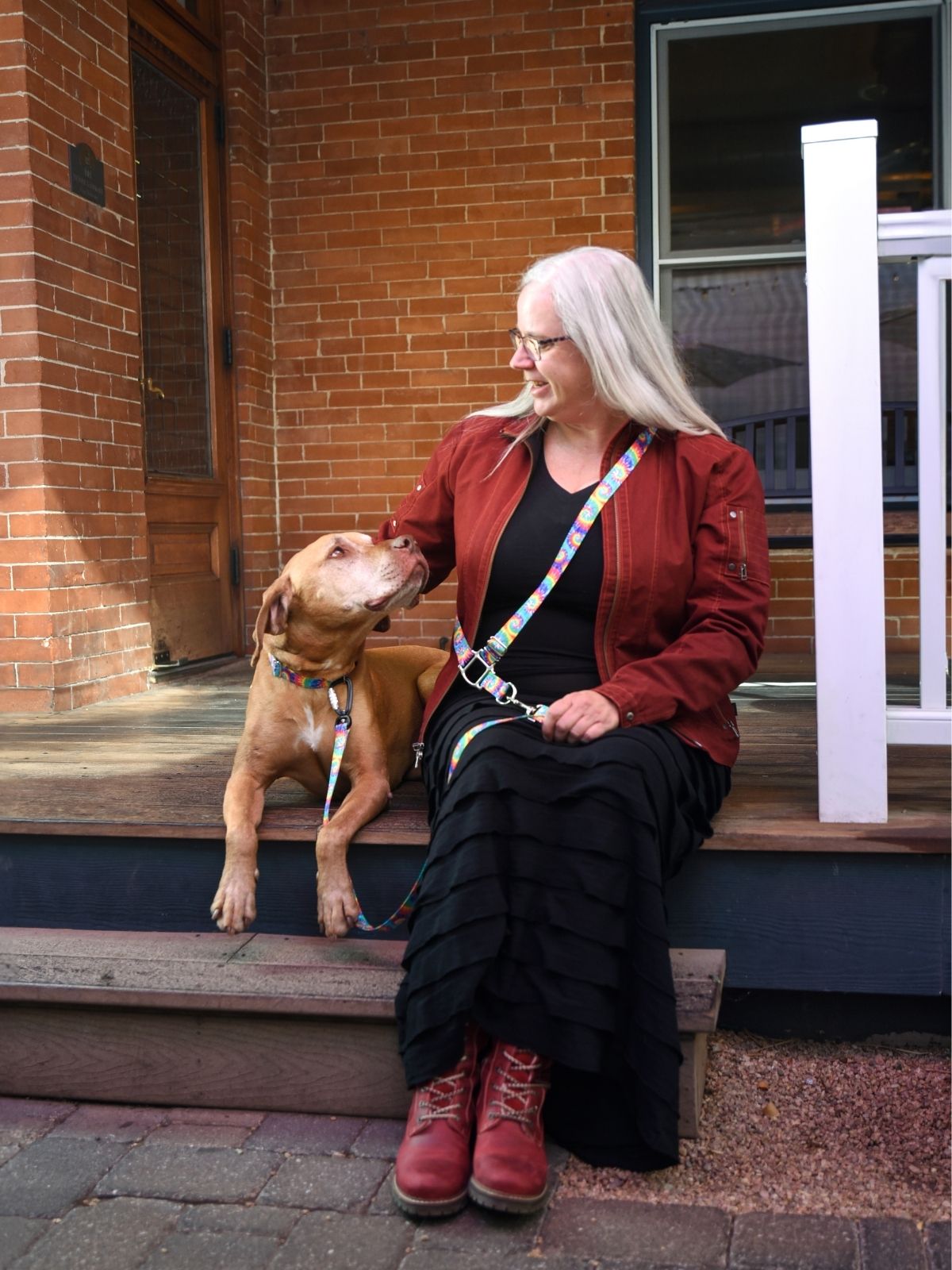 Rowan, an apricot colored dog, and his guardian are sitting on the wood steps outside a red brick home, they are looking at each other after a full day's photo shoot. Wendy is using a tie-dye pattern cross body leash and Rowan is wearing a matching collar.