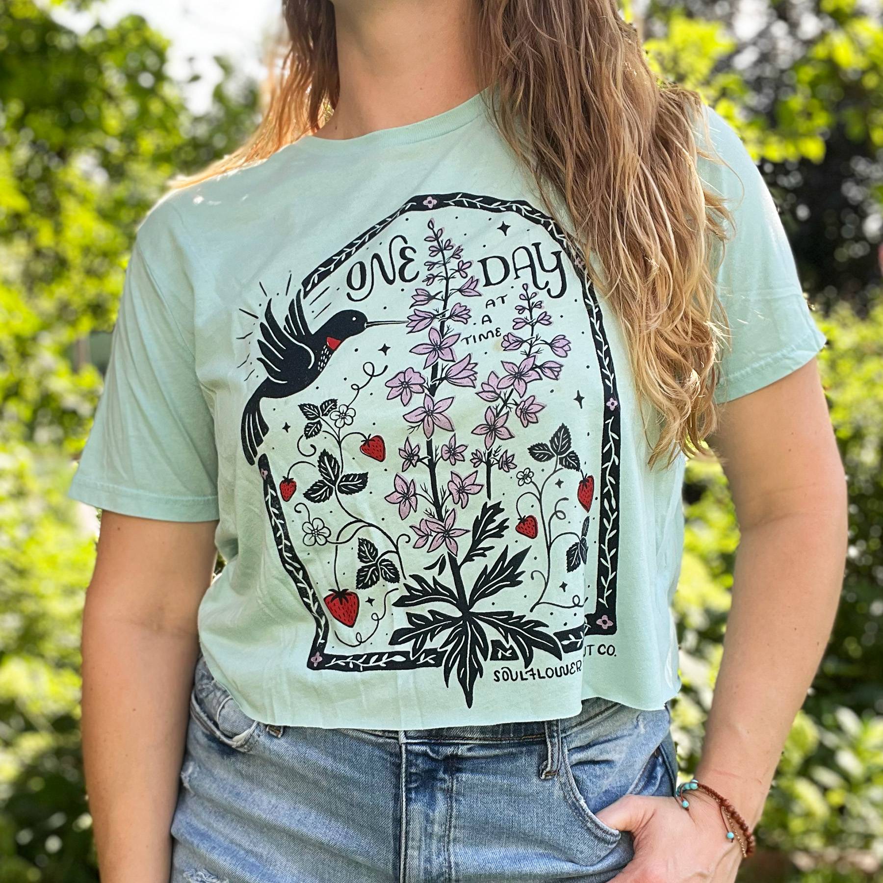 woman wearing a blue crop top featuring the July birith flower illustratoin created by Cornflower Print Co. with a hummingbird, larkspur and strawberries