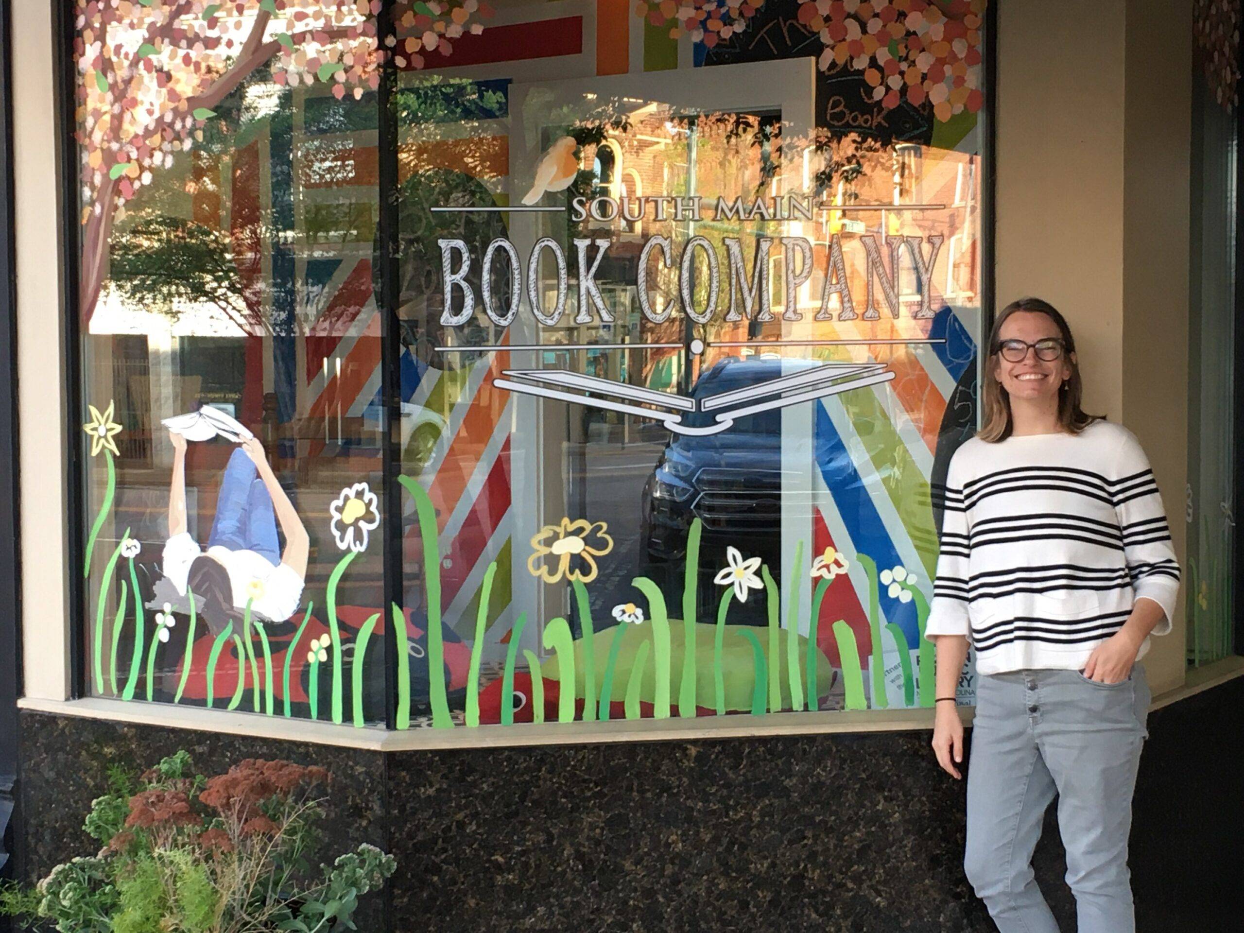 Female owner of independent bookstore stands in front of shop windows painted with the name of store - South Main Book Company - along with scene of person reading a book in flowers and vibrant green grass