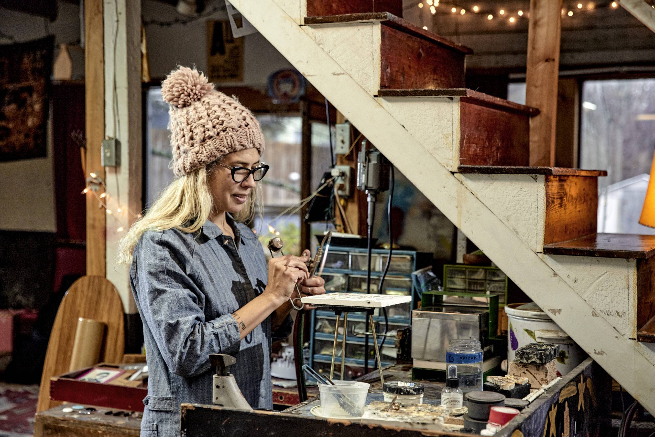 woman wearing a knit cap, dark framed glasses, and blue coveralls standing at a waist high jewelers work station, preparing to light an acetylene torch