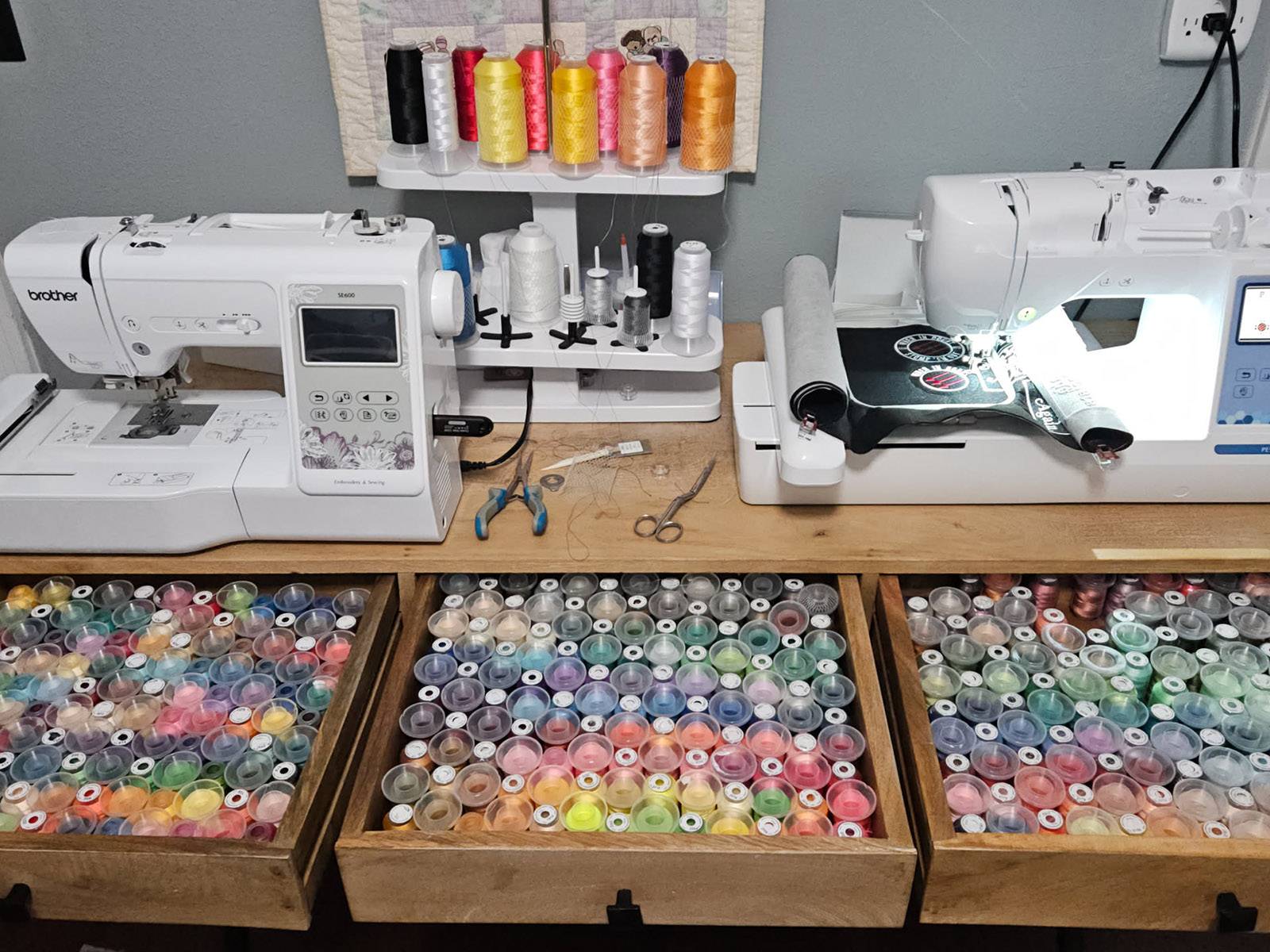 An image of two embroidery machines; a PE900 and a SE600, sitting on a desk, with drawers pulled out from the desk displaying a wide range of different colored embroidery threads. The PE900 is stitching out a circular patch that reads 