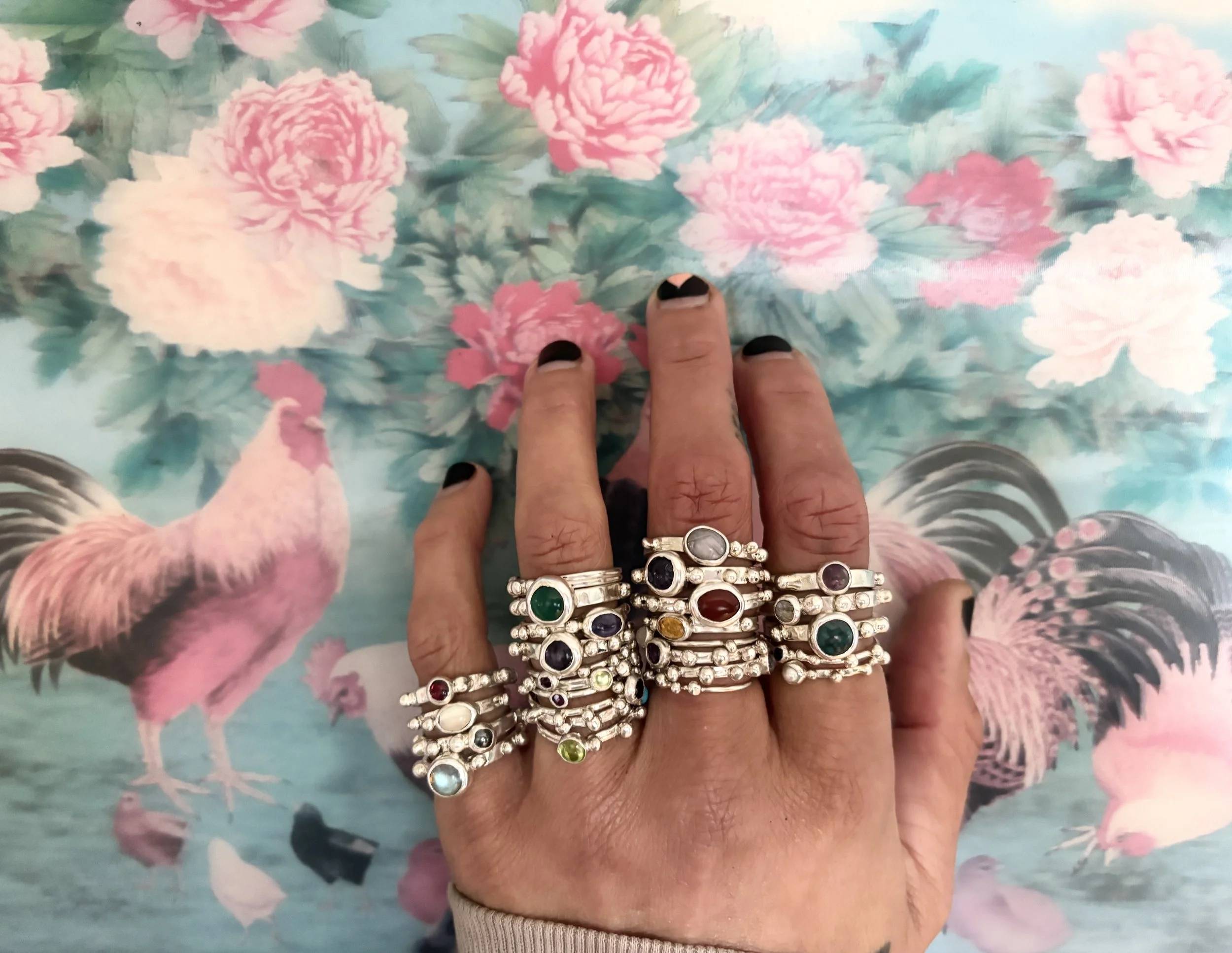 the artist's hand wearing stacking rings on all fingers except for the thumb. several rings of various gemstones on each finger. hand photo'd in front of a lenticular image of roosters and peonies.