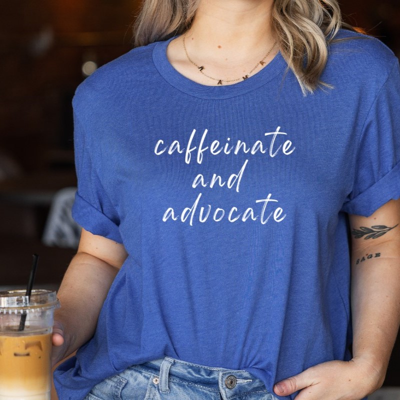 Woman wearing a blue T-shirt reading “caffeinate and advocate,” holding an iced coffee in a café setting.