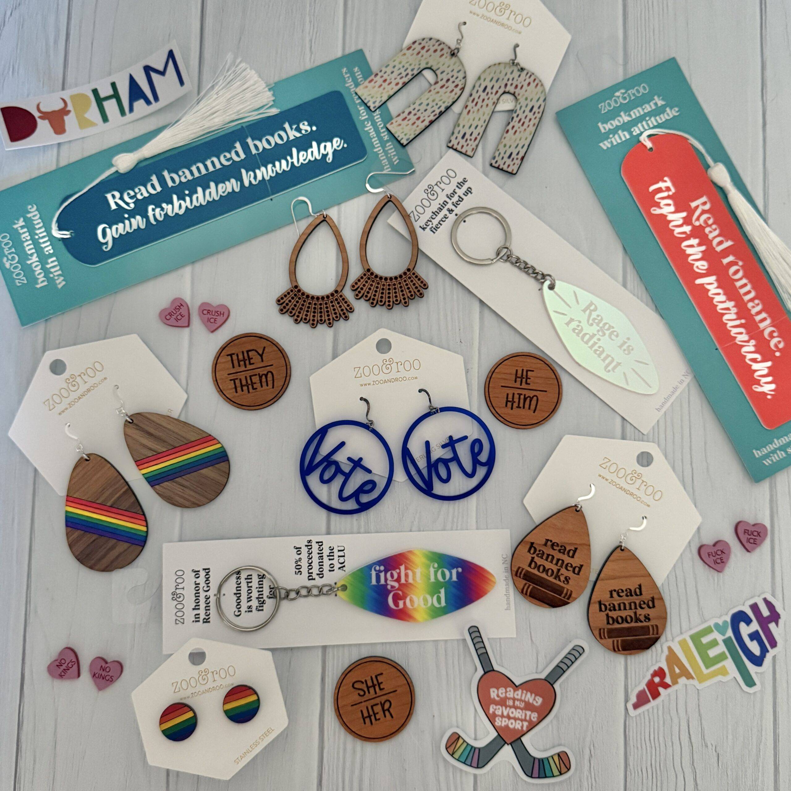 Flat lay of zoo&roo’s Fierce Collection including VOTE earrings, pride rainbow earrings, Read Banned Books bookmark, Rage is Radiant keychain, Read Romance Fight the Patriarchy bookmark, pronoun earrings, and Fight for Good keychain on a white background.