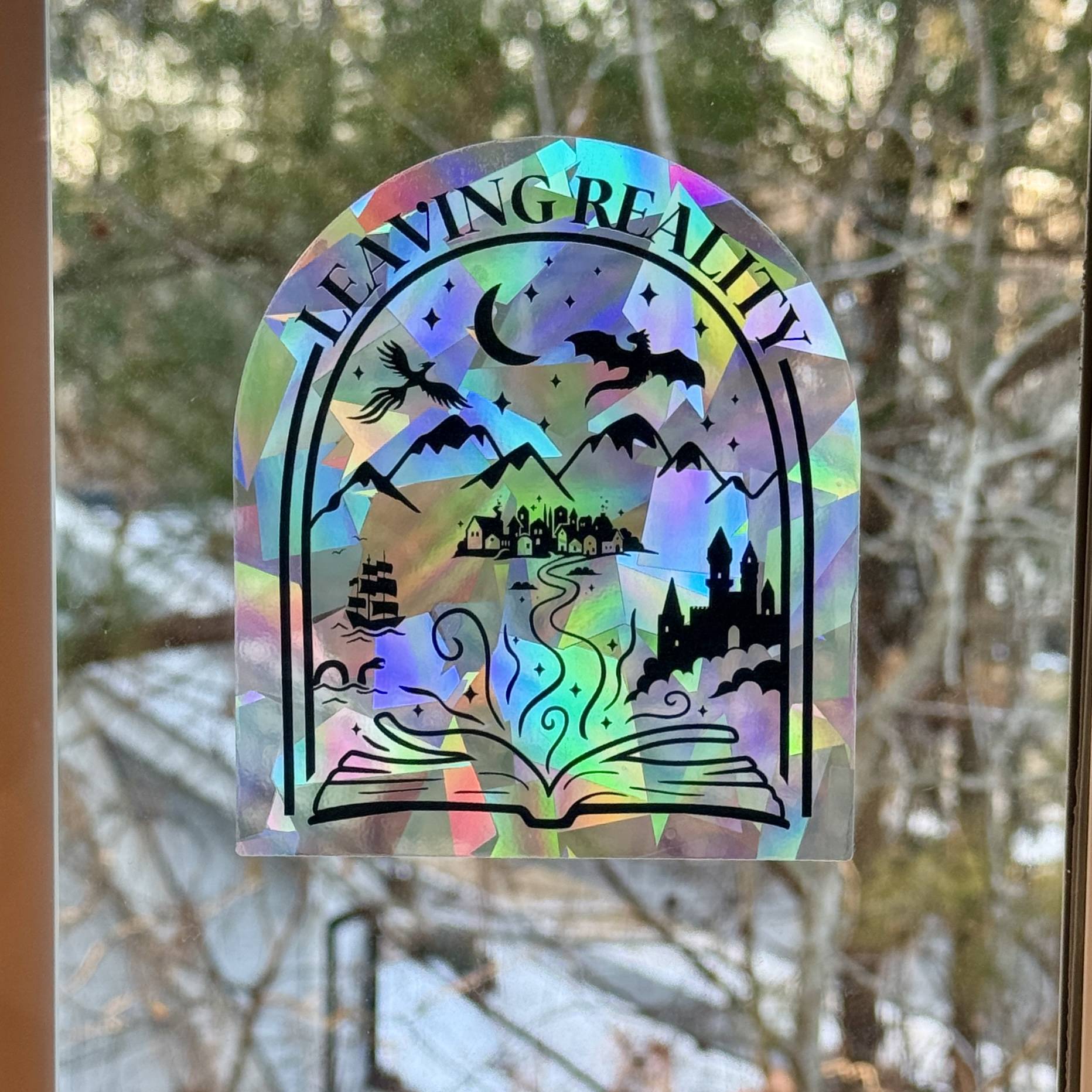Holographic “Leaving Reality” suncatcher by zoo&roo displayed in a window, featuring an open book with fantasy worlds emerging from its pages including dragons, a castle, mountains, and a tall ship, casting rainbow light.