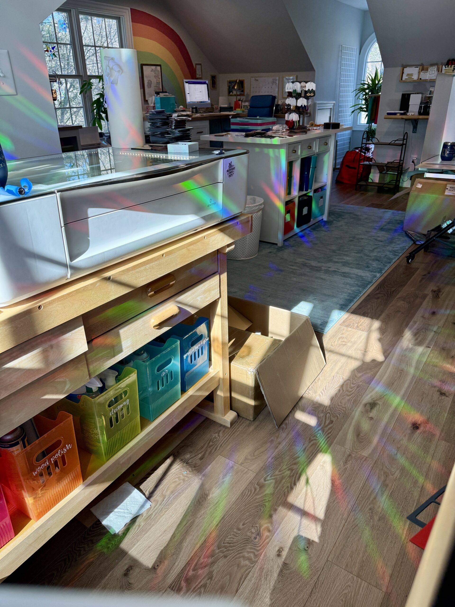 The zoo&roo studio in North Carolina with rainbow light casting across the floors and laser engraver from holographic suncatchers hanging in the windows, with a painted rainbow mural visible on the wall.