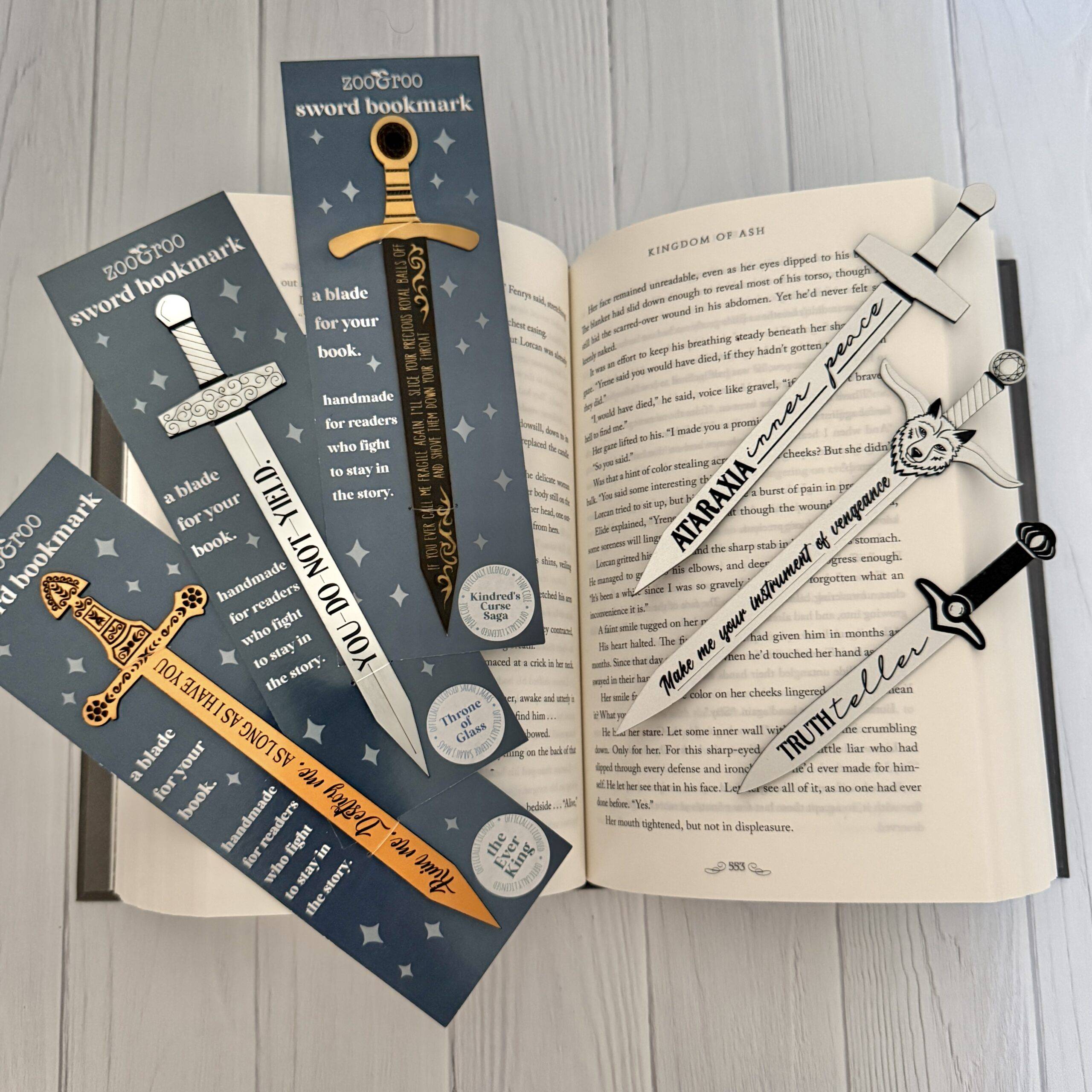 Six handmade sword bookmarks from zoo&roo fanned across an open copy of Kingdom of Ash, including Ataraxia (ACOTAR), You Do Not Yield (Throne of Glass), Ruin Me Destroy Me (Ever King), Royal Balls (Kindred’s Curse Saga), and Make Me Your Instrument of Vengeance (Dire Bound).